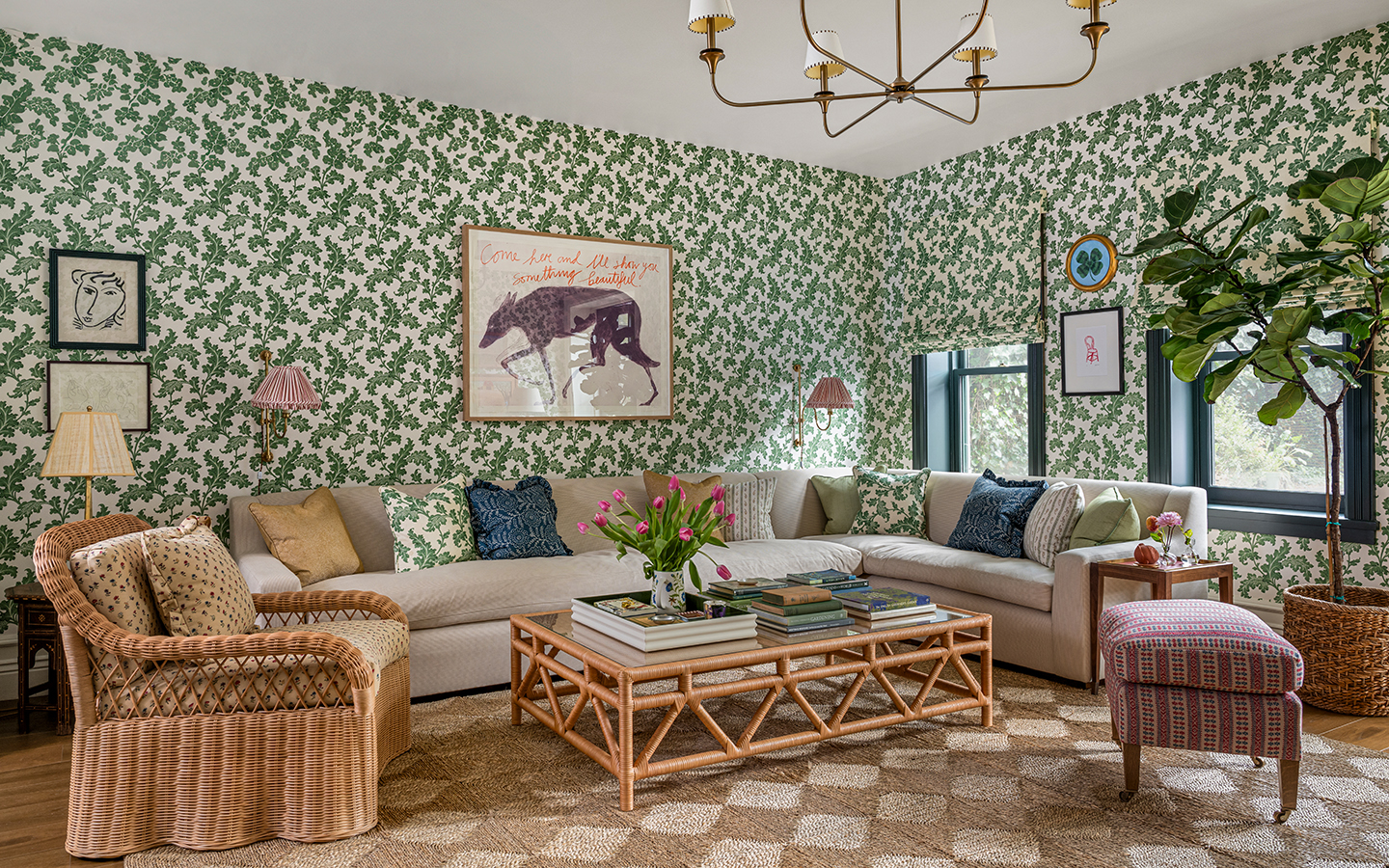 A living room with green floral wallpaper and matching blinds. A mutli-arm chandelier hangs above a bamboo glass-topped coffee table, rattan armchair, neutral corner sofa and patterned footstool. A large jute diamond pattern rug covers the floor