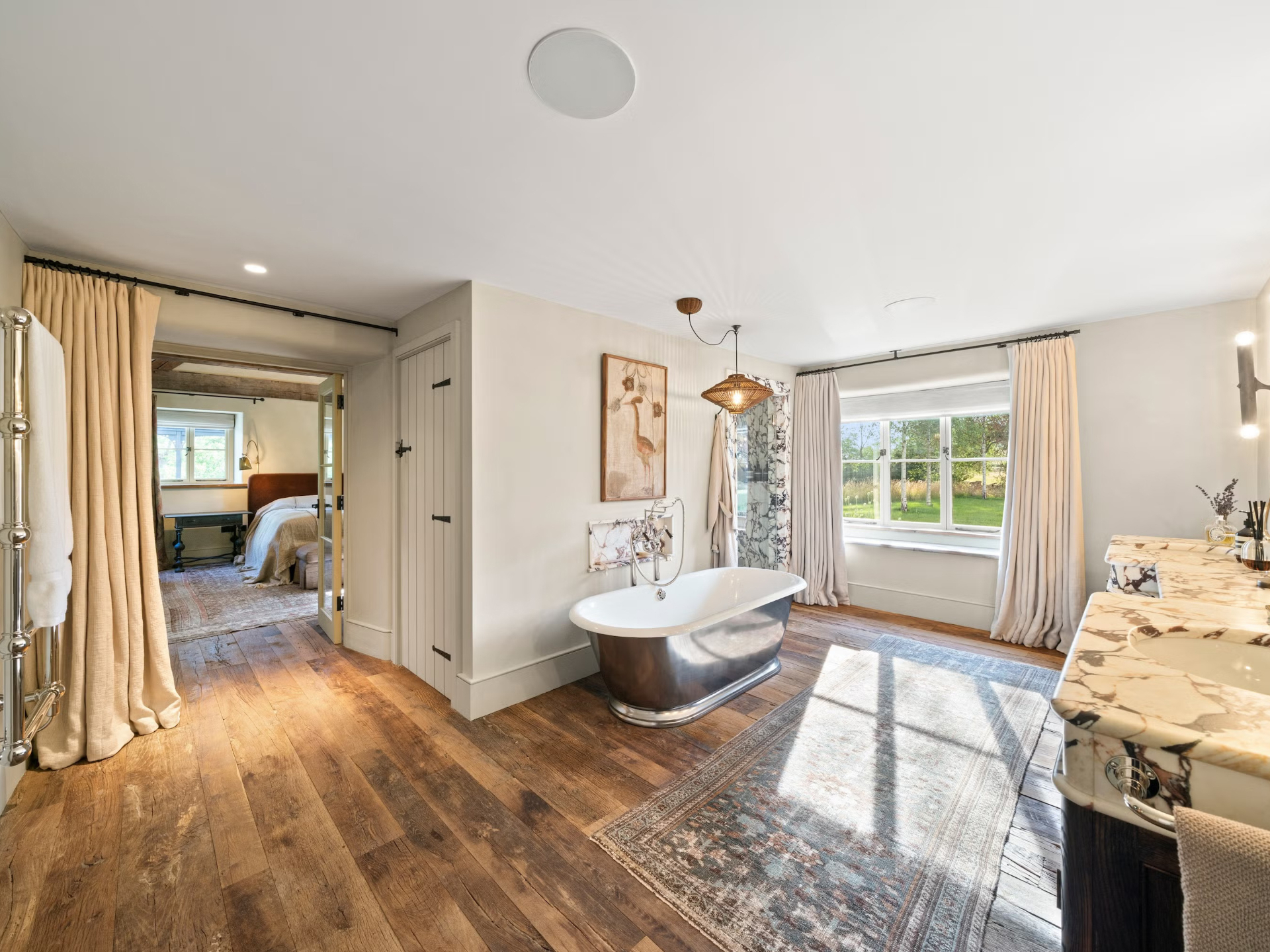 a country bathroom with wooden flooring, a silver freestanding bath tub, marble sink vanity units adn a large window with long white curtains