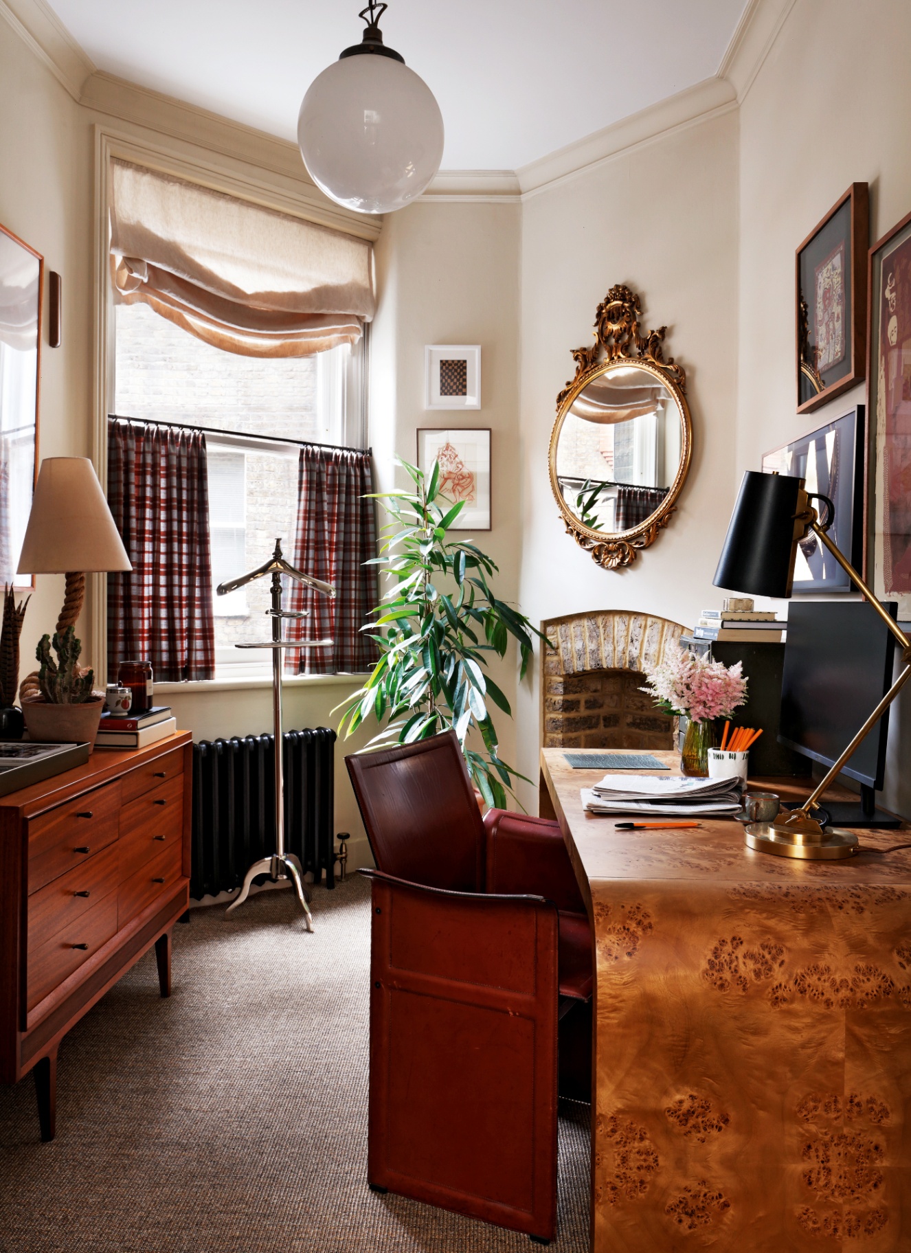 Light-filled bay window workspace featuring a figure-grained burl wood desk, vintage mid-century teak sideboard, burgundy leather armchair, and ornate gold mirror.