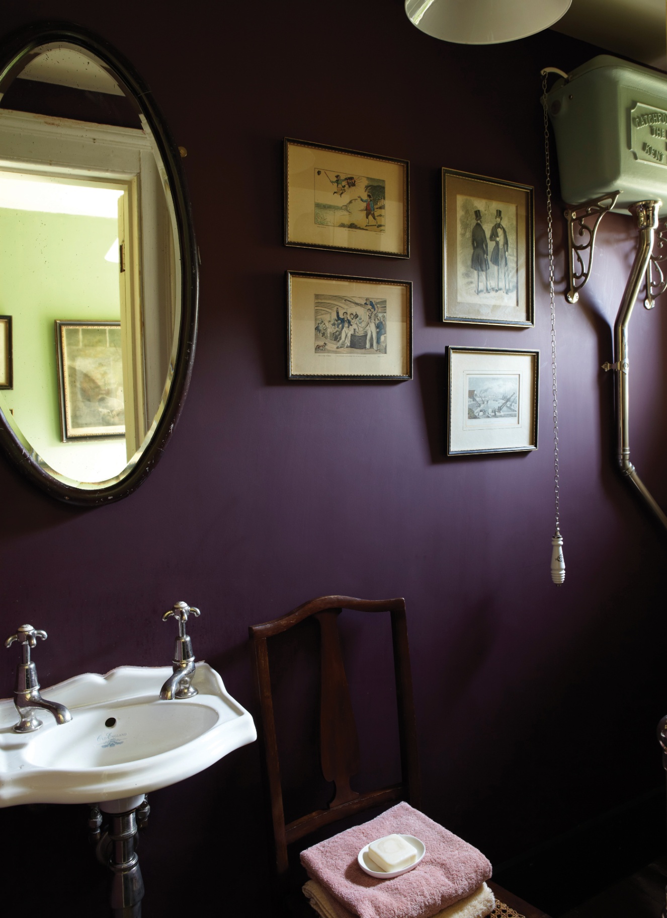 Dark plum bathroom featuring a pedestal sink with silver fixtures, an oval mirror, and framed vintage artwork on deep purple walls. A high-level toilet cistern with a pull chain sits beside a wooden chair holding folded towels