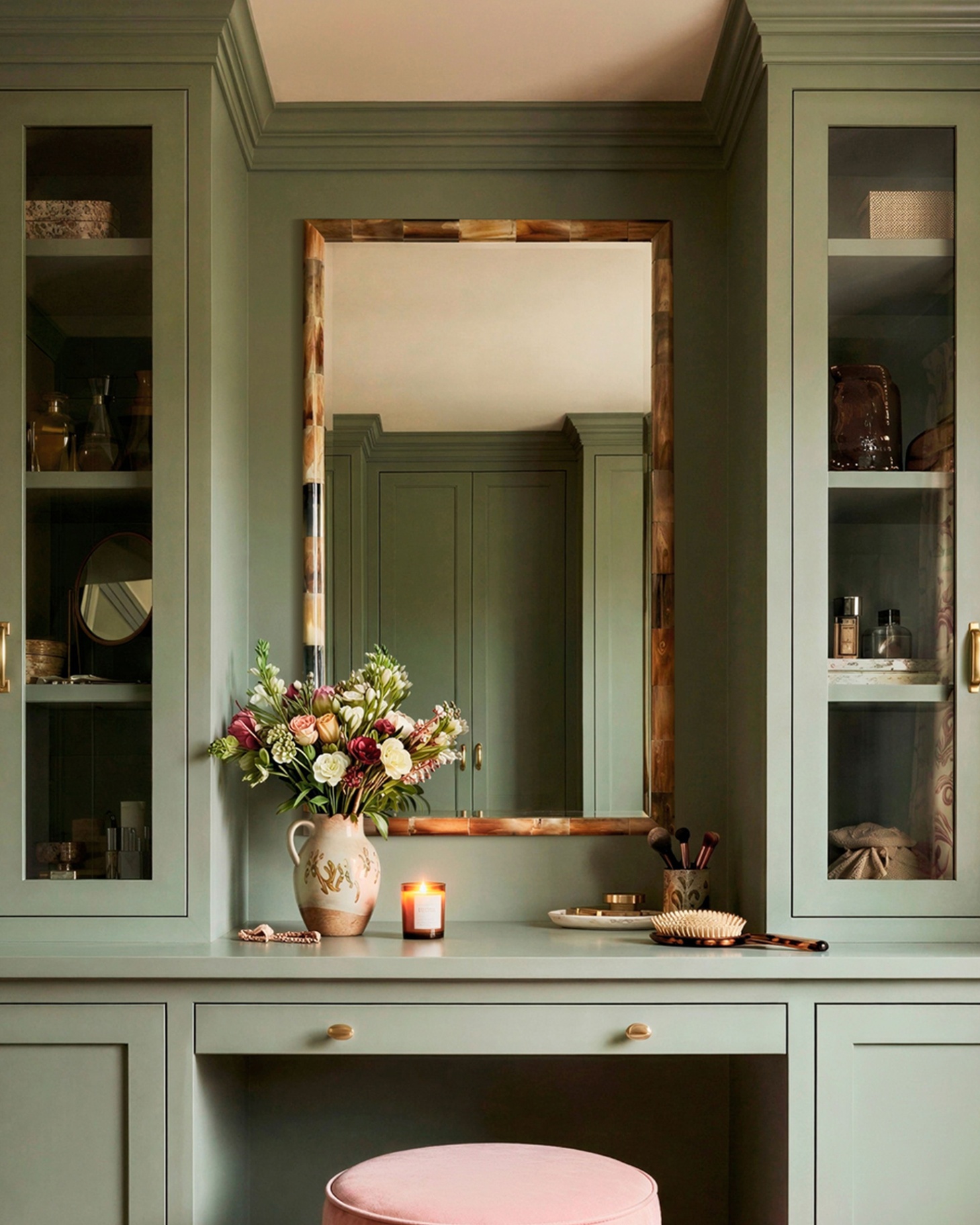 green built in dressing table area, with tall built in cabinets either side, a large rectangle tortoise shell mirror, a vase of flowers and a small pink stool