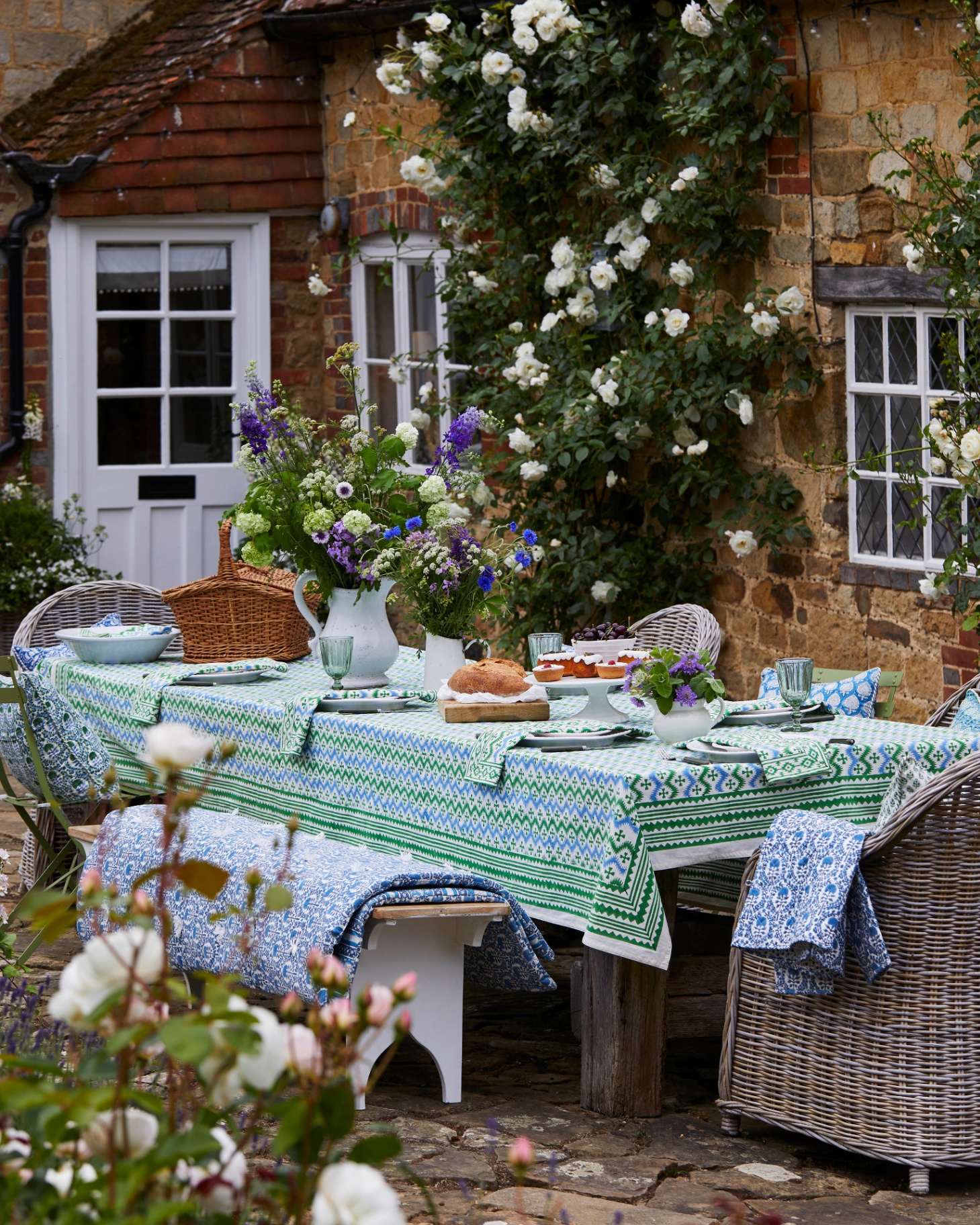 an outdoor dining table with a blue, white and green patterned tablecloth, rattan tub chairs with matching cushions and a table full of flowers and party food