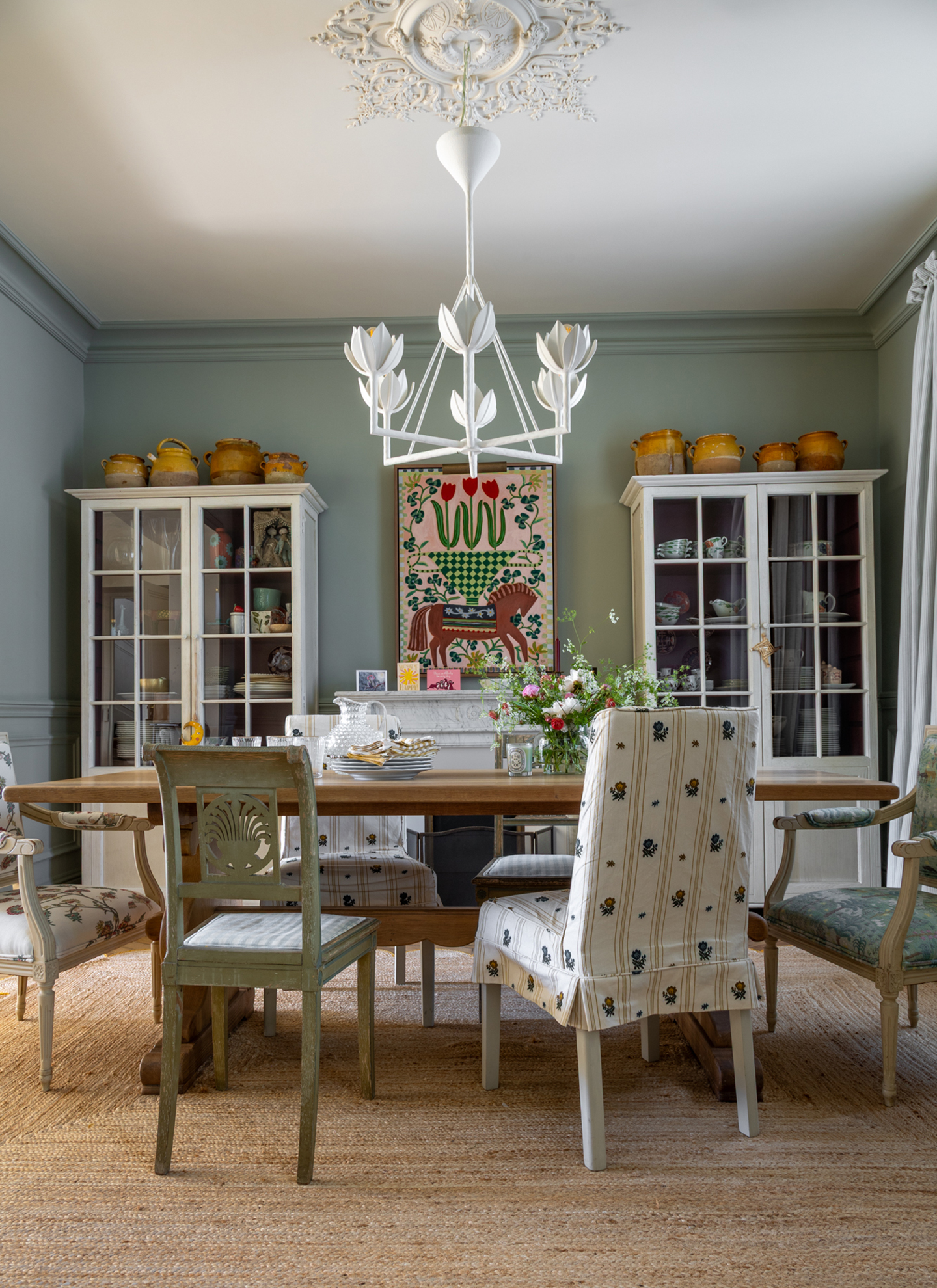 Elegant dining room featuring a white tulip-shaped chandelier, a wooden table with mixed chairs, and twin display cabinets against sage walls