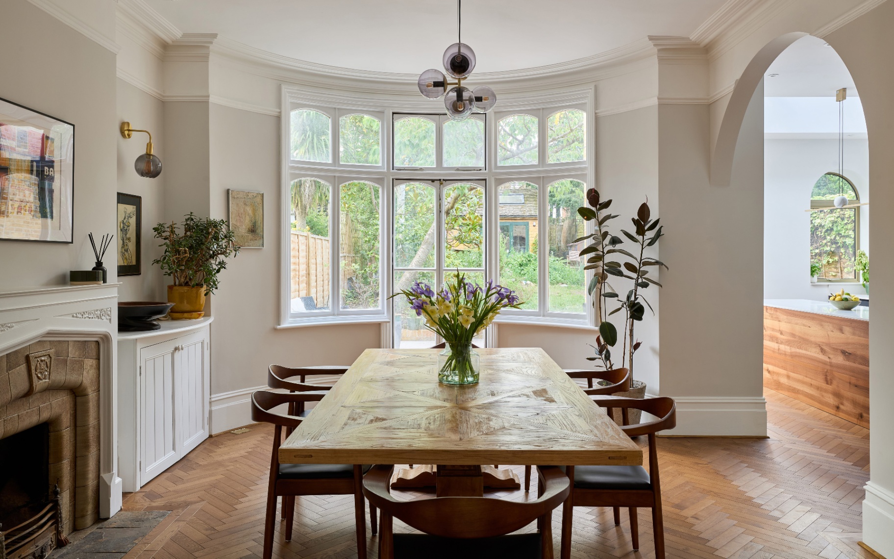 a dining room with wooden parquet flooring, a dining table with 5 wooden chairs and a vase filled with flowers, a large curved window, a fireplace to the left and a white built in cupboard next to it