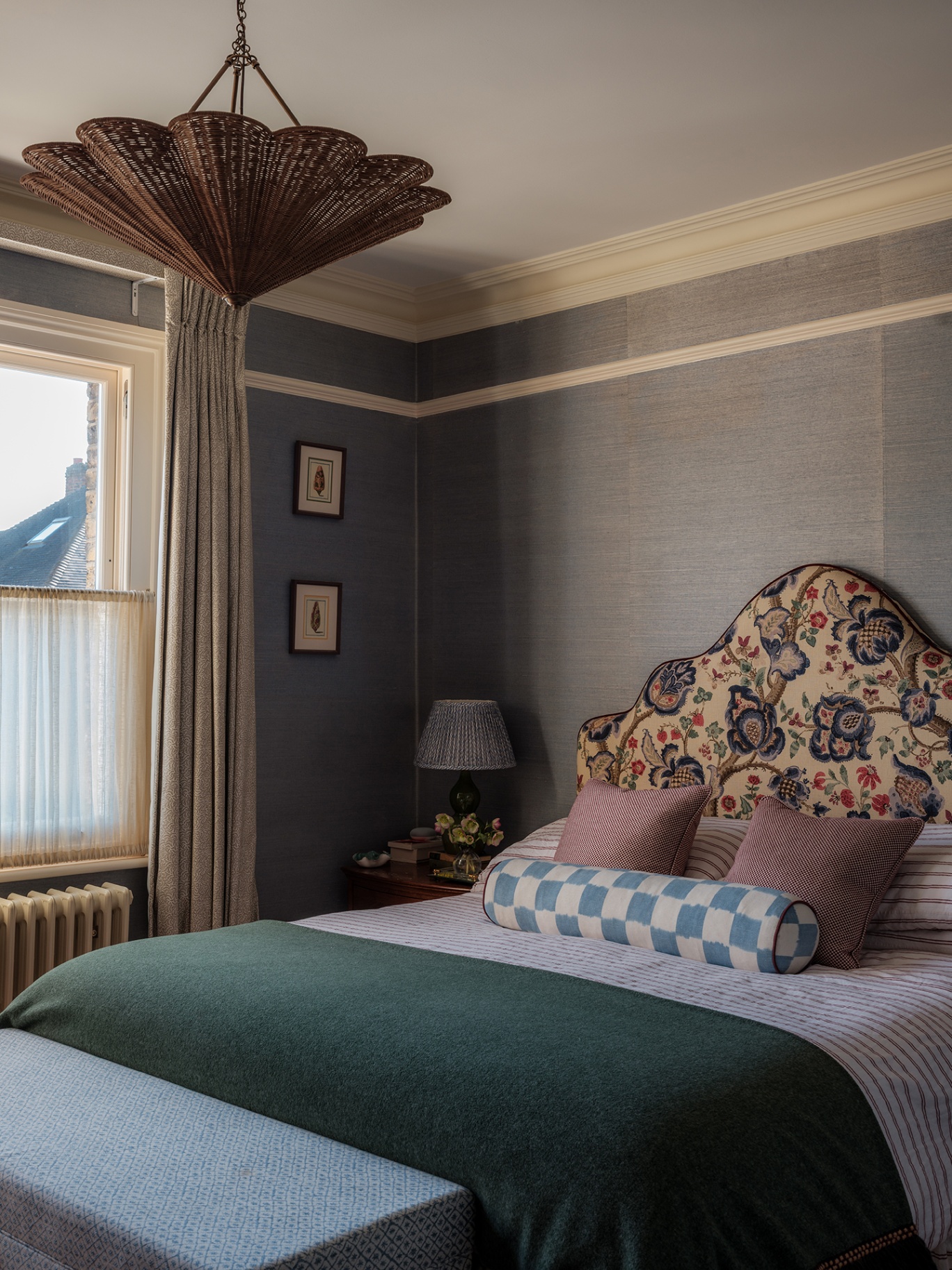 A bedroom with blue hemp covered walls and a double bed with a statement upholstered headboard. A large rattan pendant hangs above and to the left a window is dressed with curtains and a cafe curtain