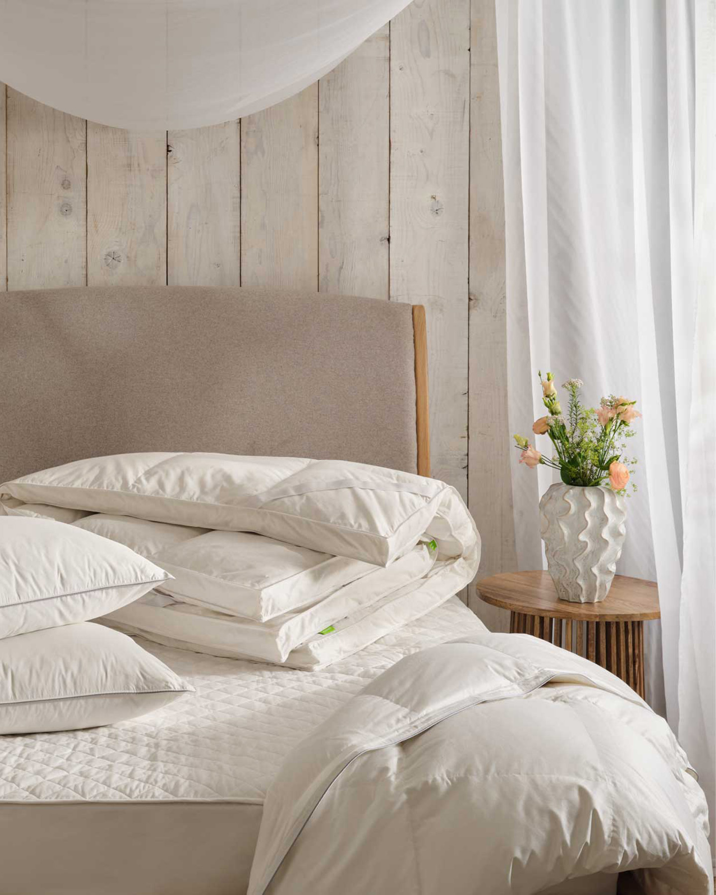 a folded duvet and mattress protector on a bed with a wooden bedside table text to it with a white vase and flowers