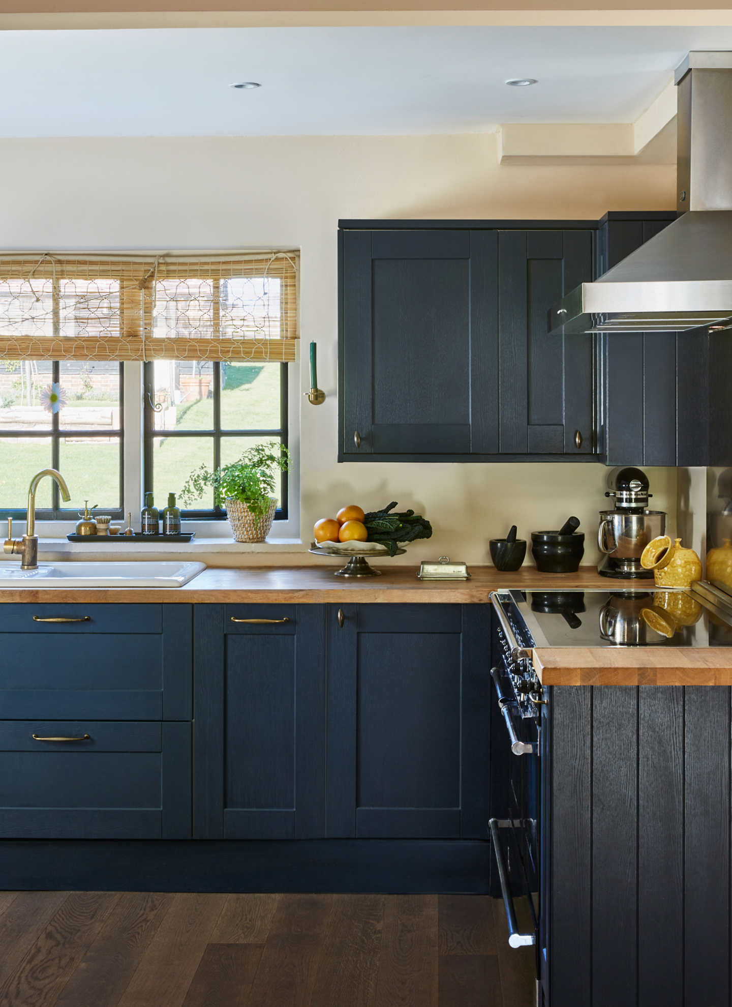 a kitchen with dark blue cabinets, a window with bamboo blinds and a tray on the window ledge with soap and dish brushes