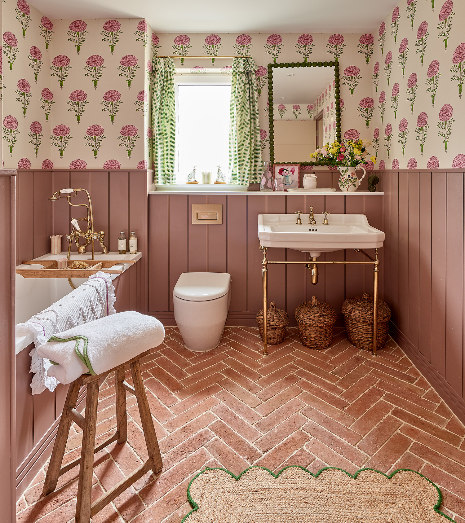 ca pietra bathroom with flagstone flooring, pink half panelled walls with cream and pink rose block print wallpaper on top, a small jute scallope rug, a traditional basin with a green bobin mirror above it and a window with green gingham curtains