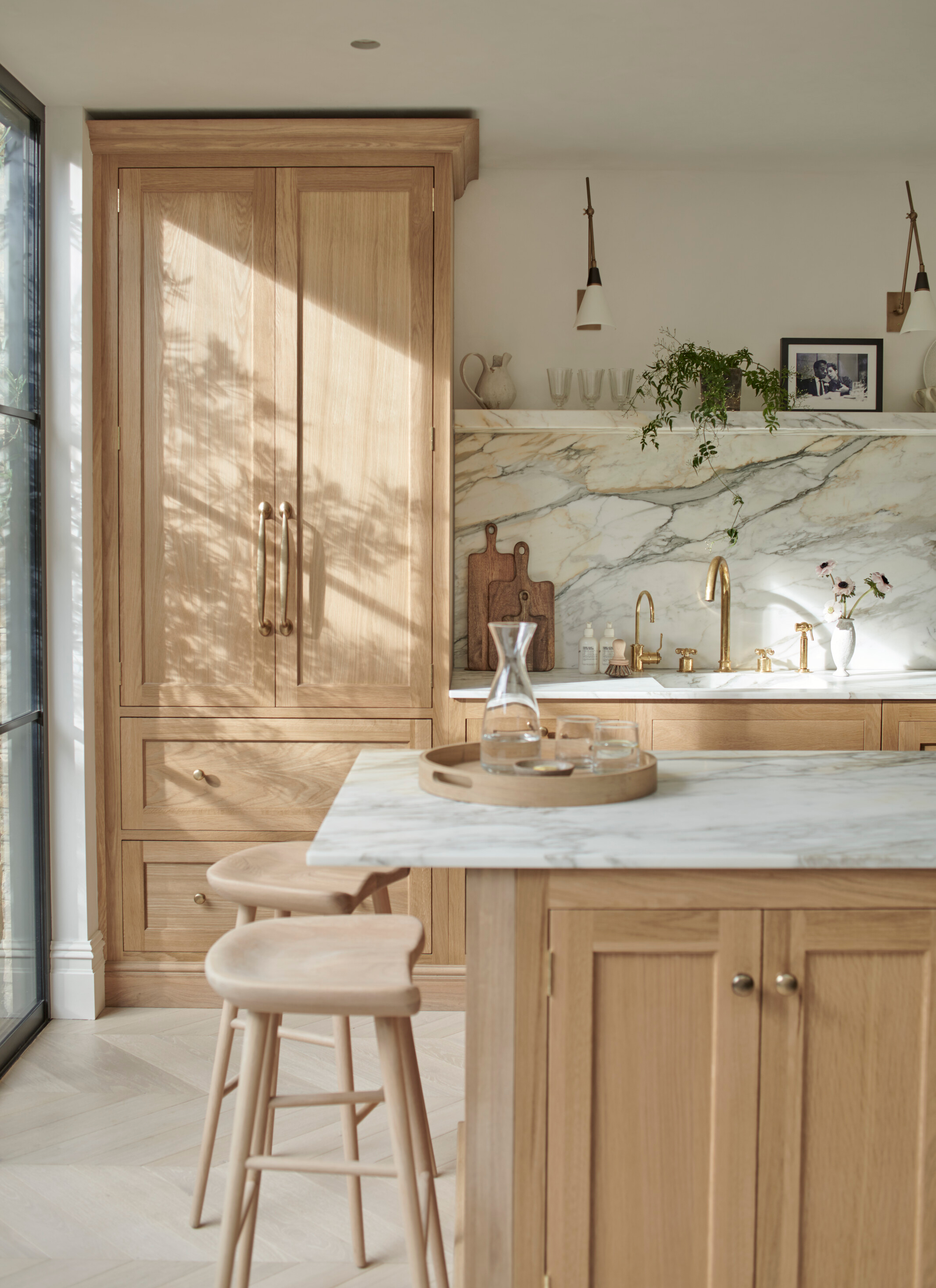 a kitchen with pale wooden cabinetry, a pale wooden kitchen island with stools and white and grey marble countertops and splashback