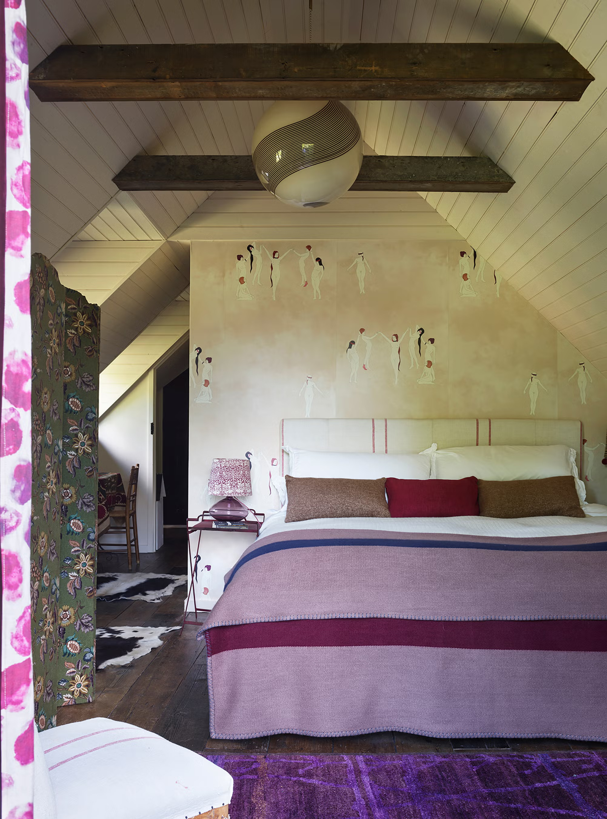 a bedroom with white walls, wooden beams, a bed with a pink throw and wallpaper with illustrations of people