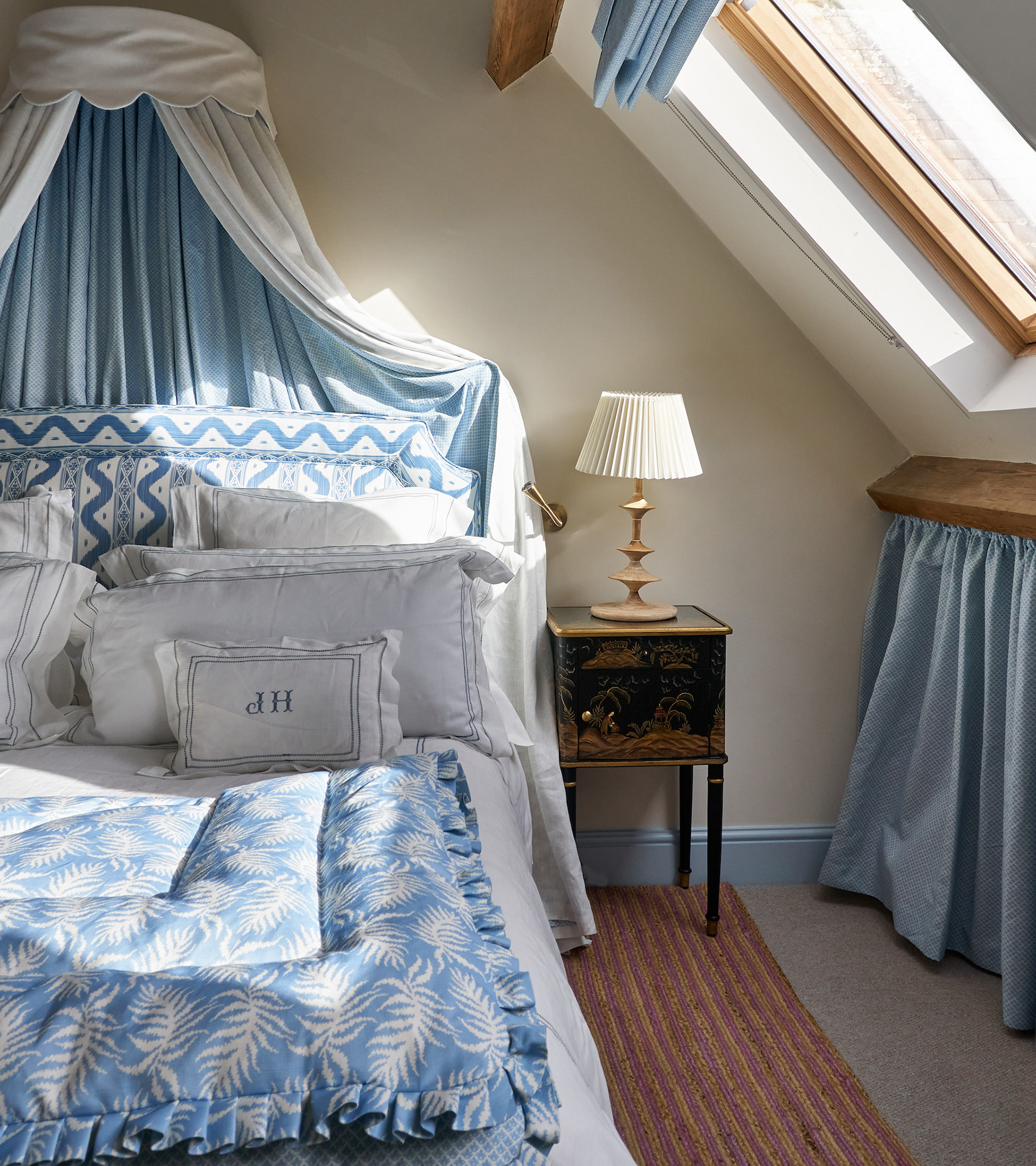 an attic bedroom with wooden beams on the ceiling, a large bed with a white canopy, a patterned blue and white headboard, a patterned blue and white bedspread with an antique wooden table and a lamp on top