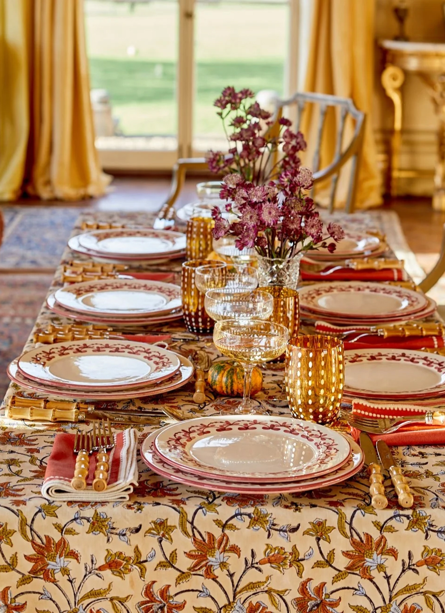 an autumn table with a cream, orange and ochre floral table cloth, bamboo cutlery, white and red patterned plates, amber tumbler with white polka dots and a vase of dark pink flowers