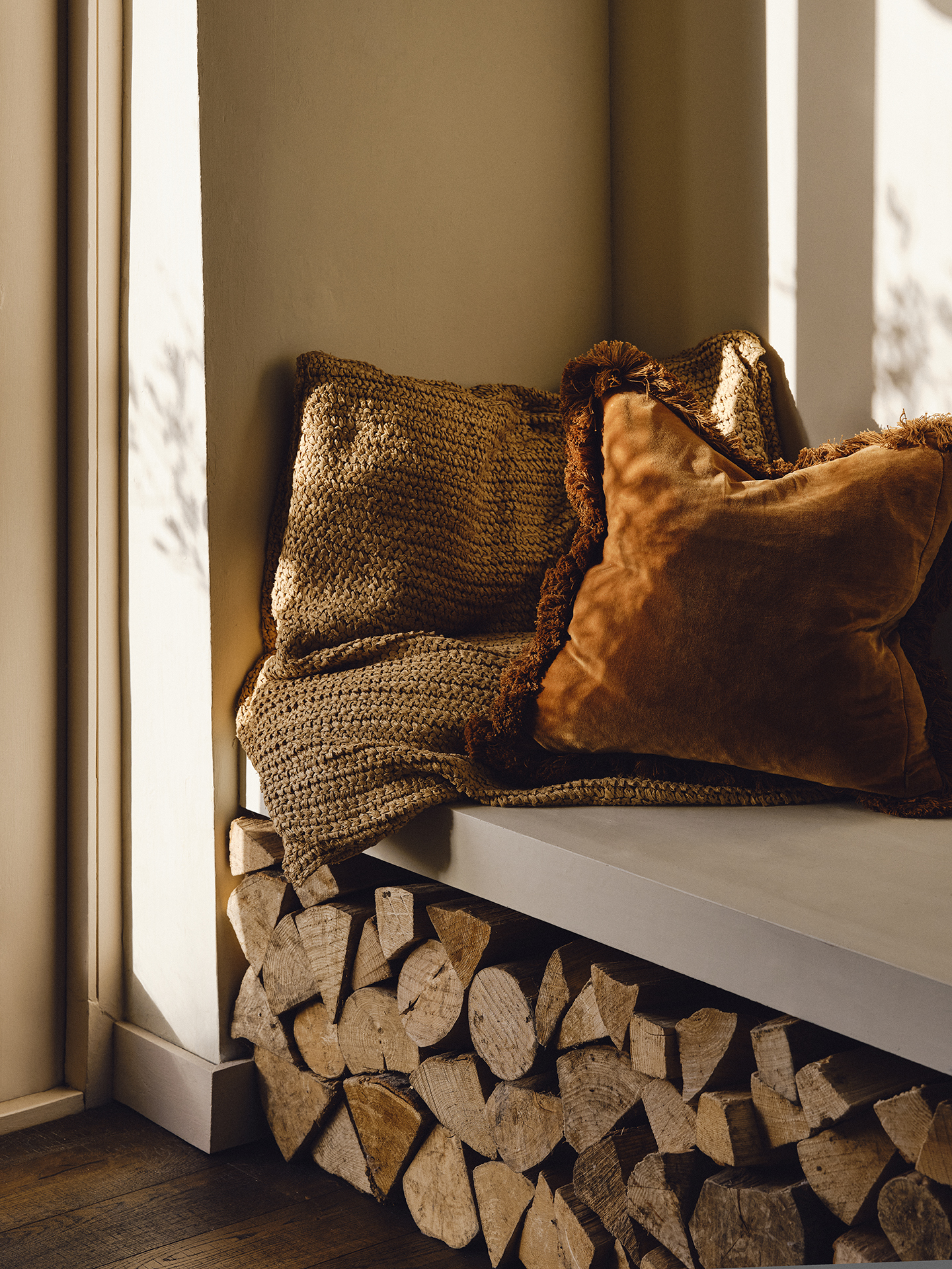 A close up shot of a woven cushion and velvet fringed cushion sat on a bench above cut wood for a fire