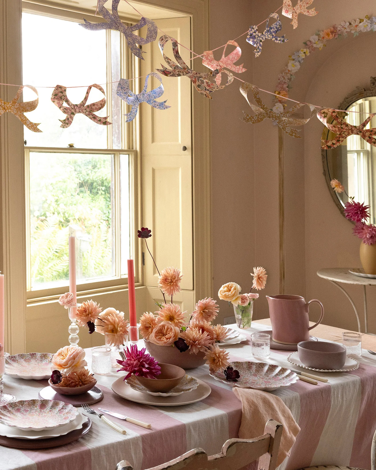a spring tablescape with a pink and white stripe table cloth, vases filled with pink flowers and liberty print floral bow bunting hanging above the table