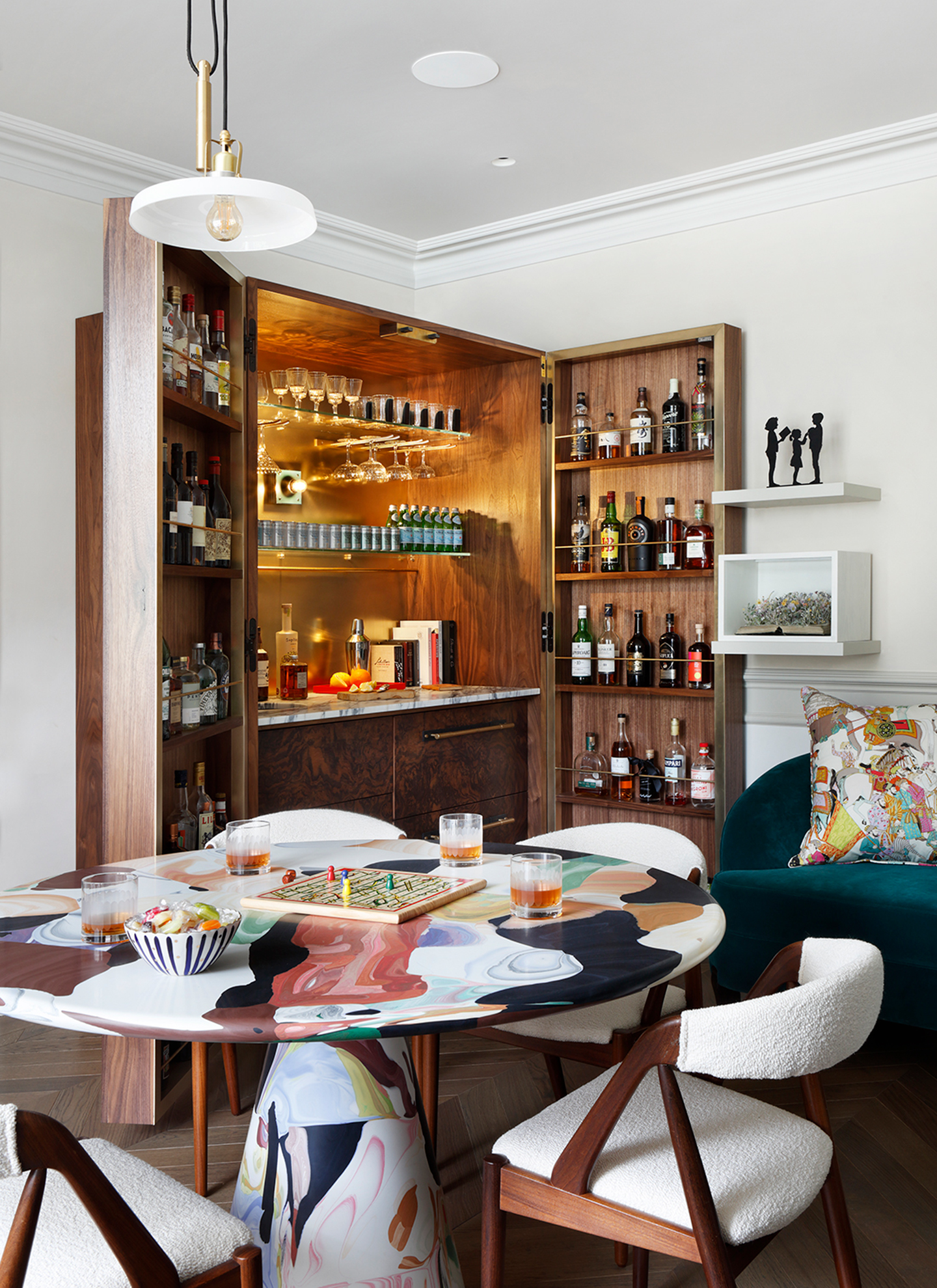A stylish room featuring an open bespoke wooden drinks cabinet with mirrored lighting, set behind a patterned round games table with board games and whitebouclé chairs.   