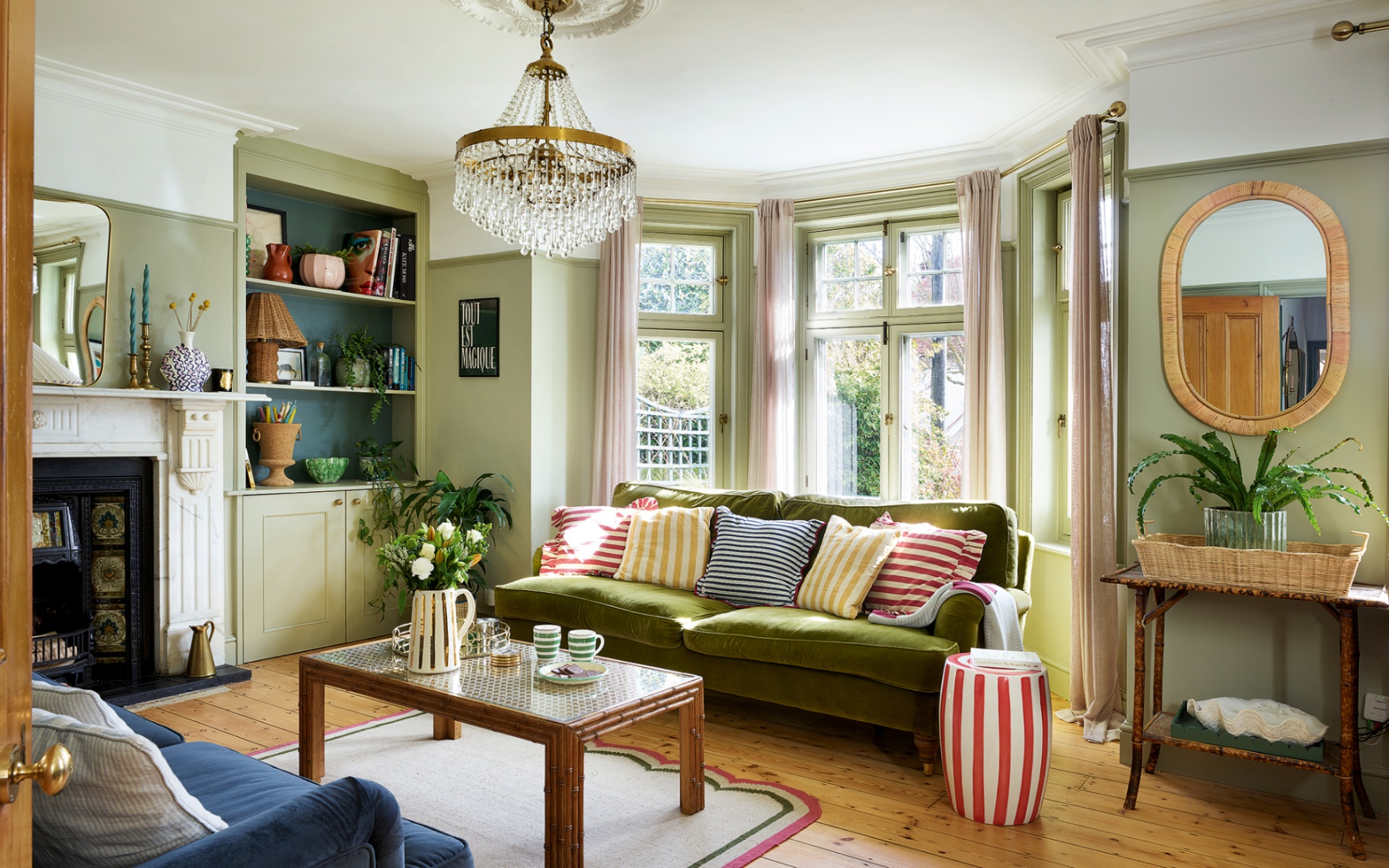 An angled shot of a living room with pale green walls, blue and green velvet sofas sat facing a vintage coffee table set atop a scalloped rug. A chandelier hangs in the middle of the room. To the left is a marble fireplace and built-in bookshelf