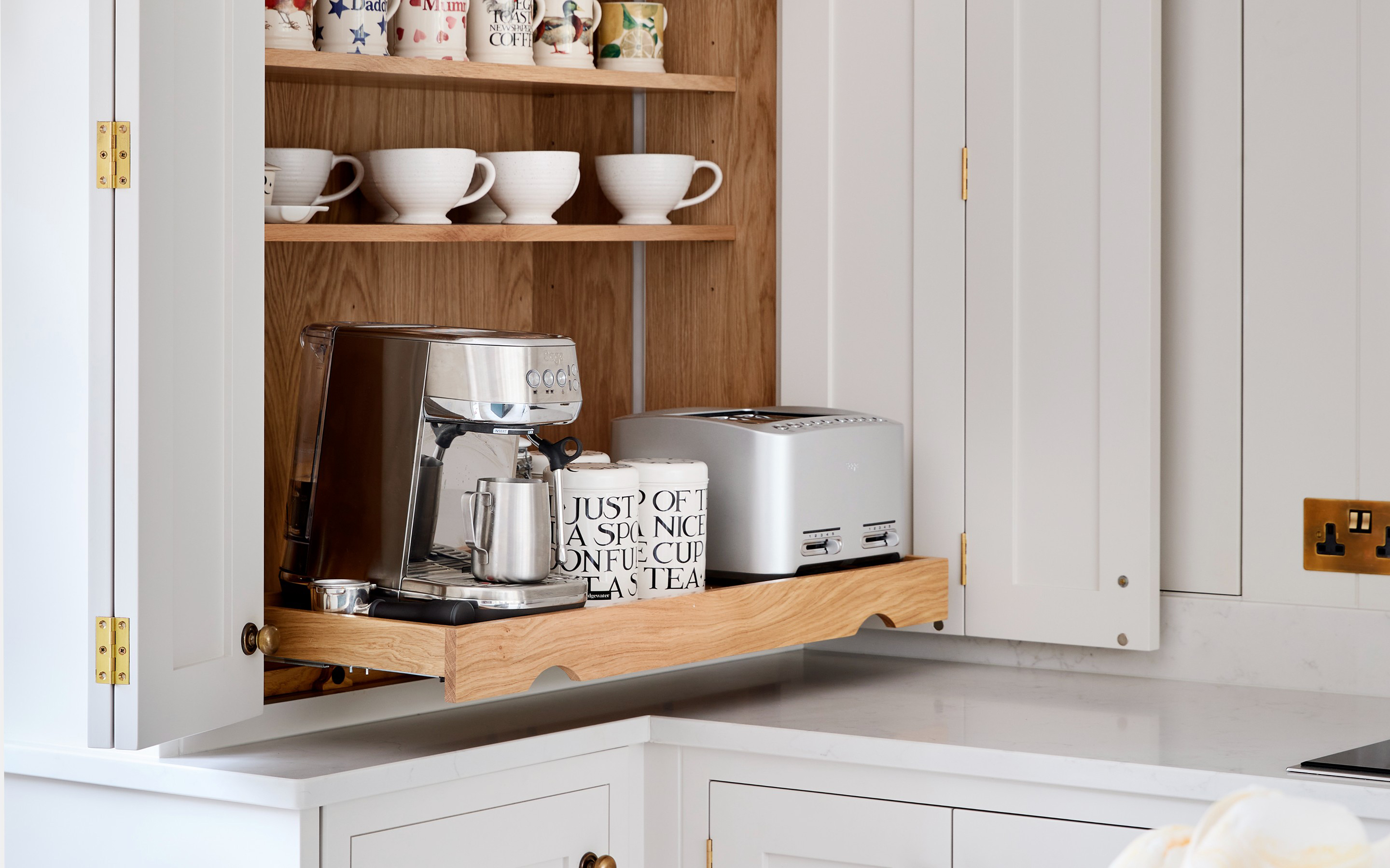 White bi-fold countertop cabinet with an extended wooden pull-out shelf holding a coffee machine and toaster, backed by oak lining and mug shelves above