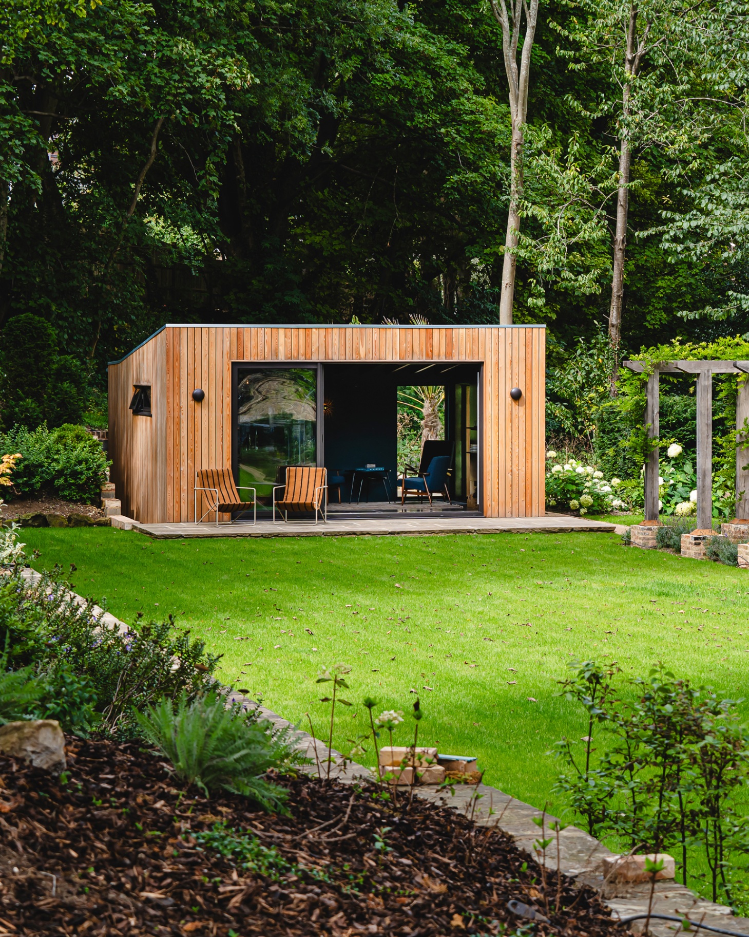 a wooden panelled garden room with two brown deck chairs outside looking out onto a green lawn with trees behind it