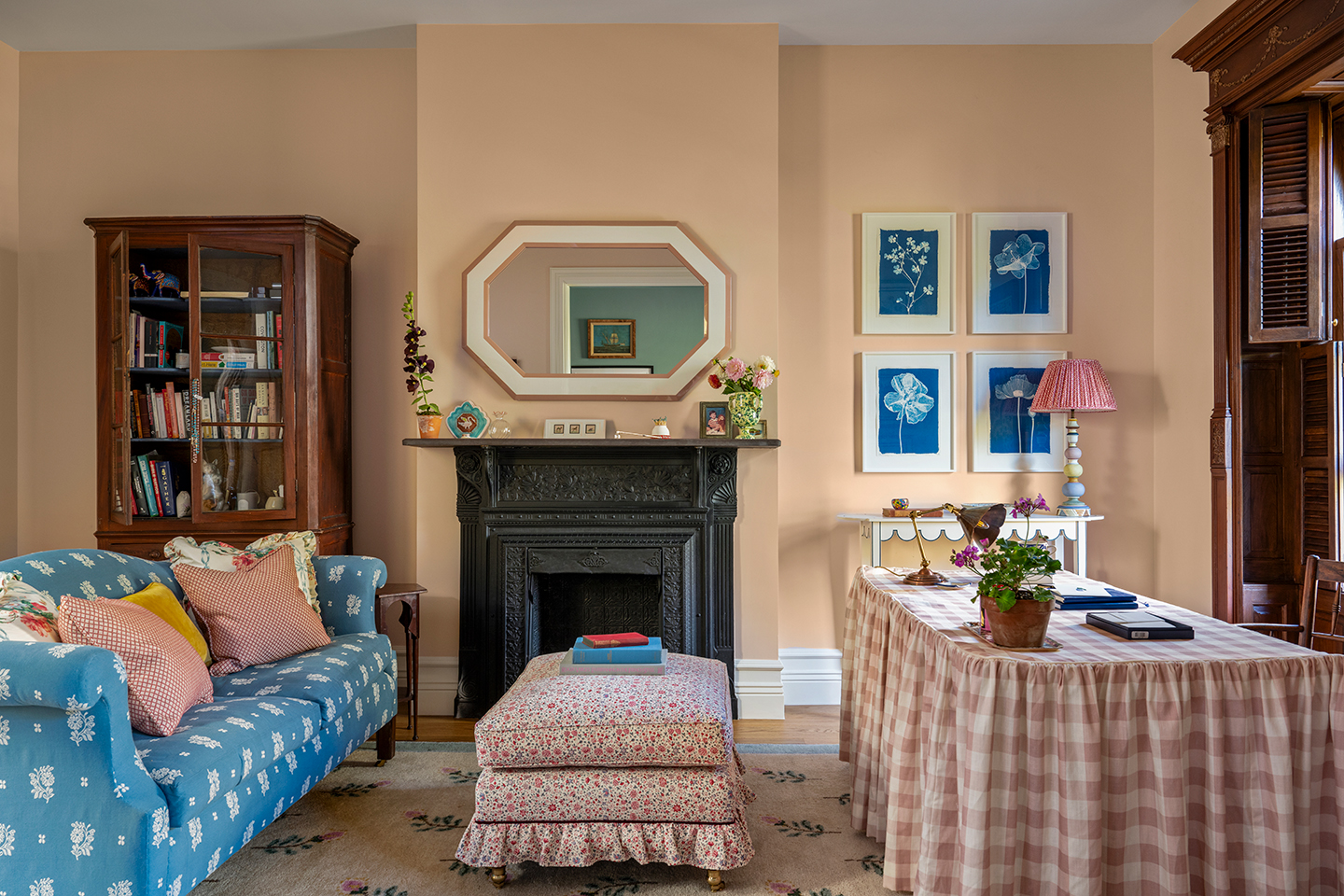 A study with a floral blue sofa, ruffled upholstered ottoman, large desk dressed with a pink check skirt. On the back wall is a wooden cabinet filled with books, an ornate fireplace with octagonal mirror above and a series of four blue botanical prints 