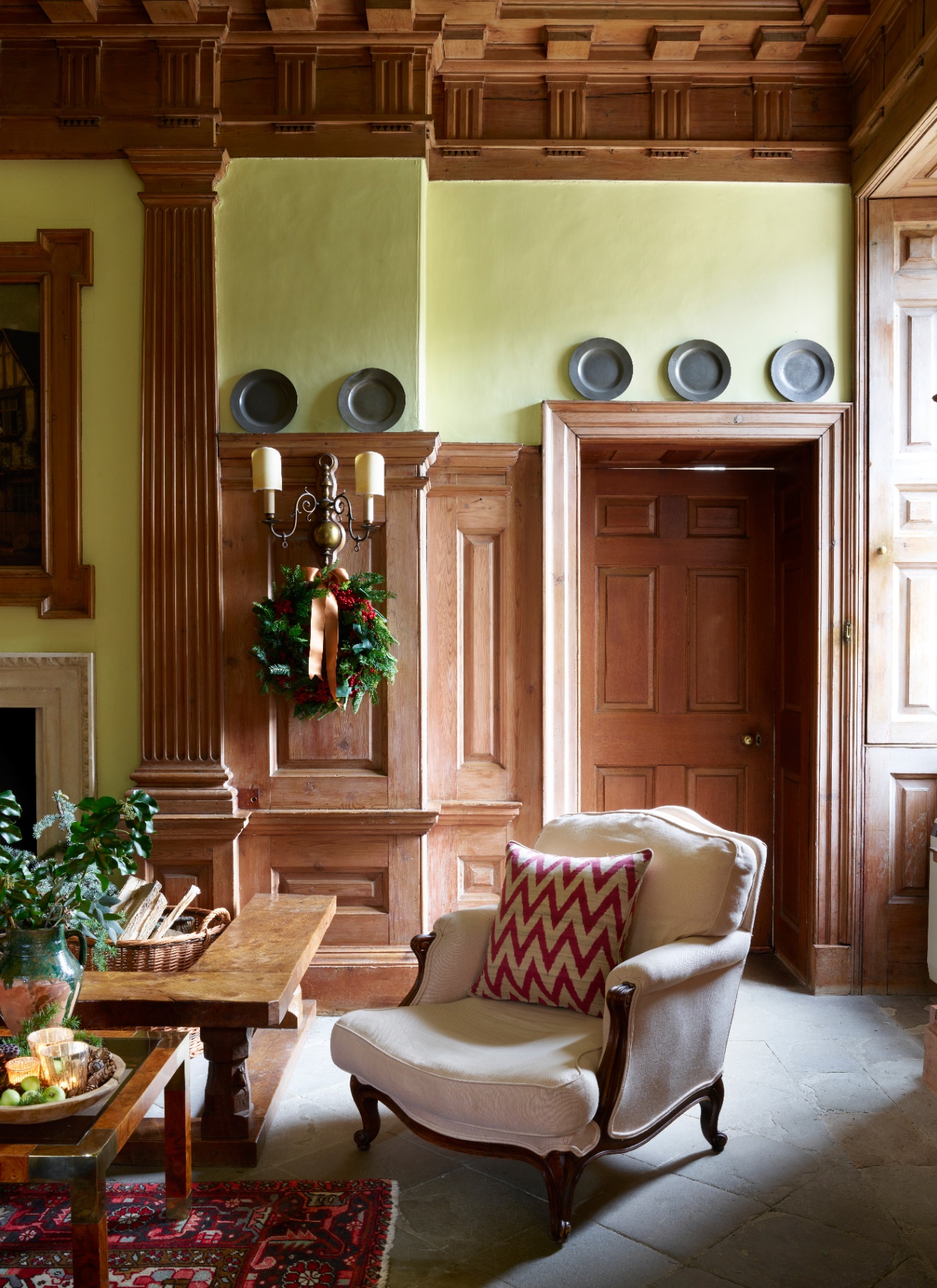 A pine and berry Christmas wreath with a caramel bow hanging from an antique brass wall sconce against wood-panelled walls in a period entrance hall