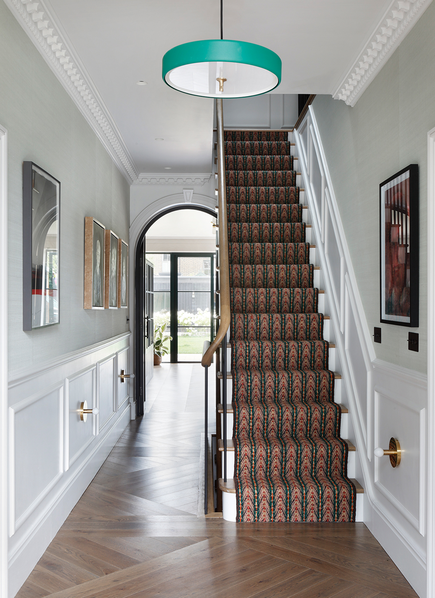 a hallway with white walls, panelling, a teal pendant shade and stairs with a red and brown patterned runner