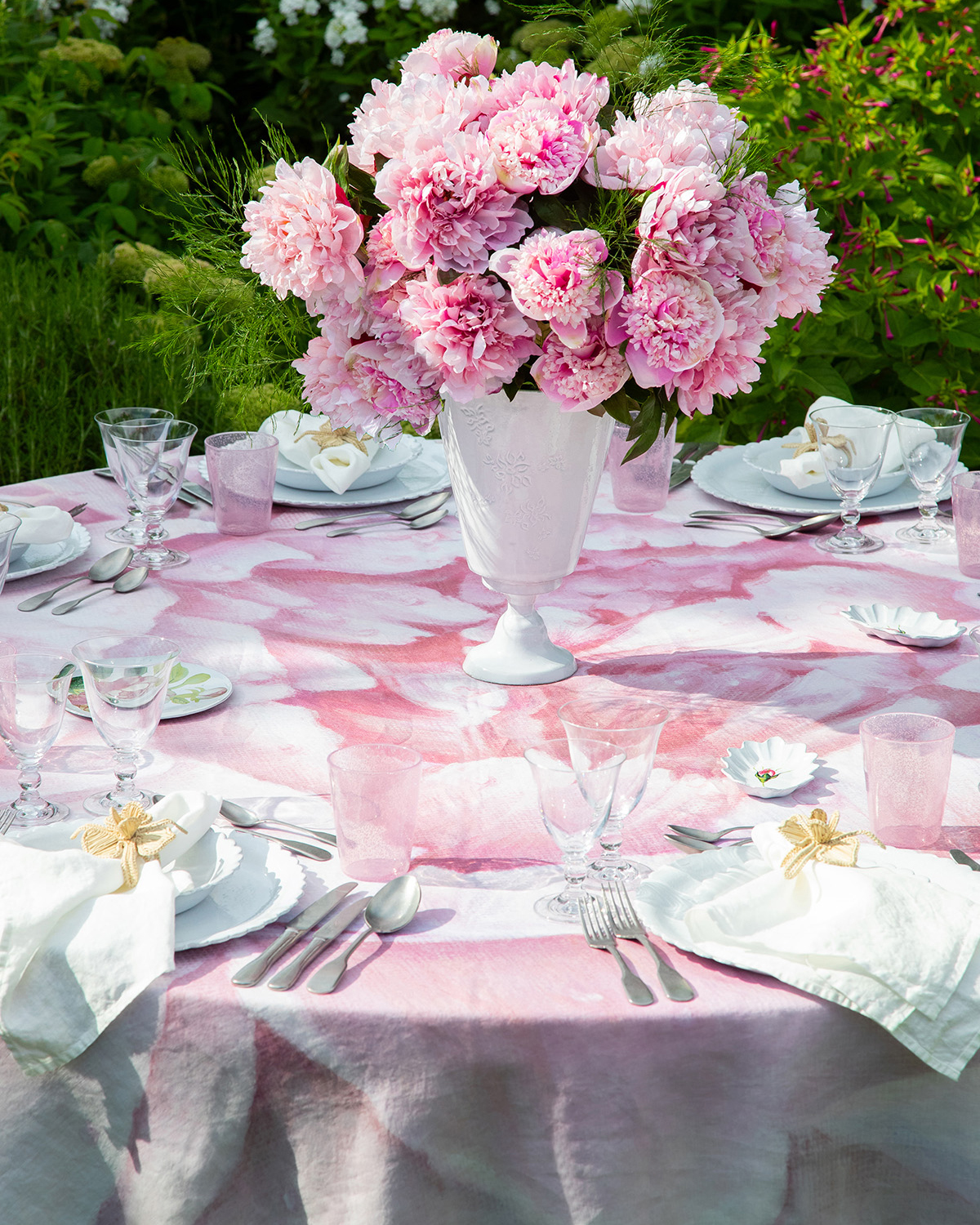 summerill and bishop pink pink table cloth on a round table, with alarge white vase in teh centre filled with pink flowers and white flower shaped plates and bowls on the table