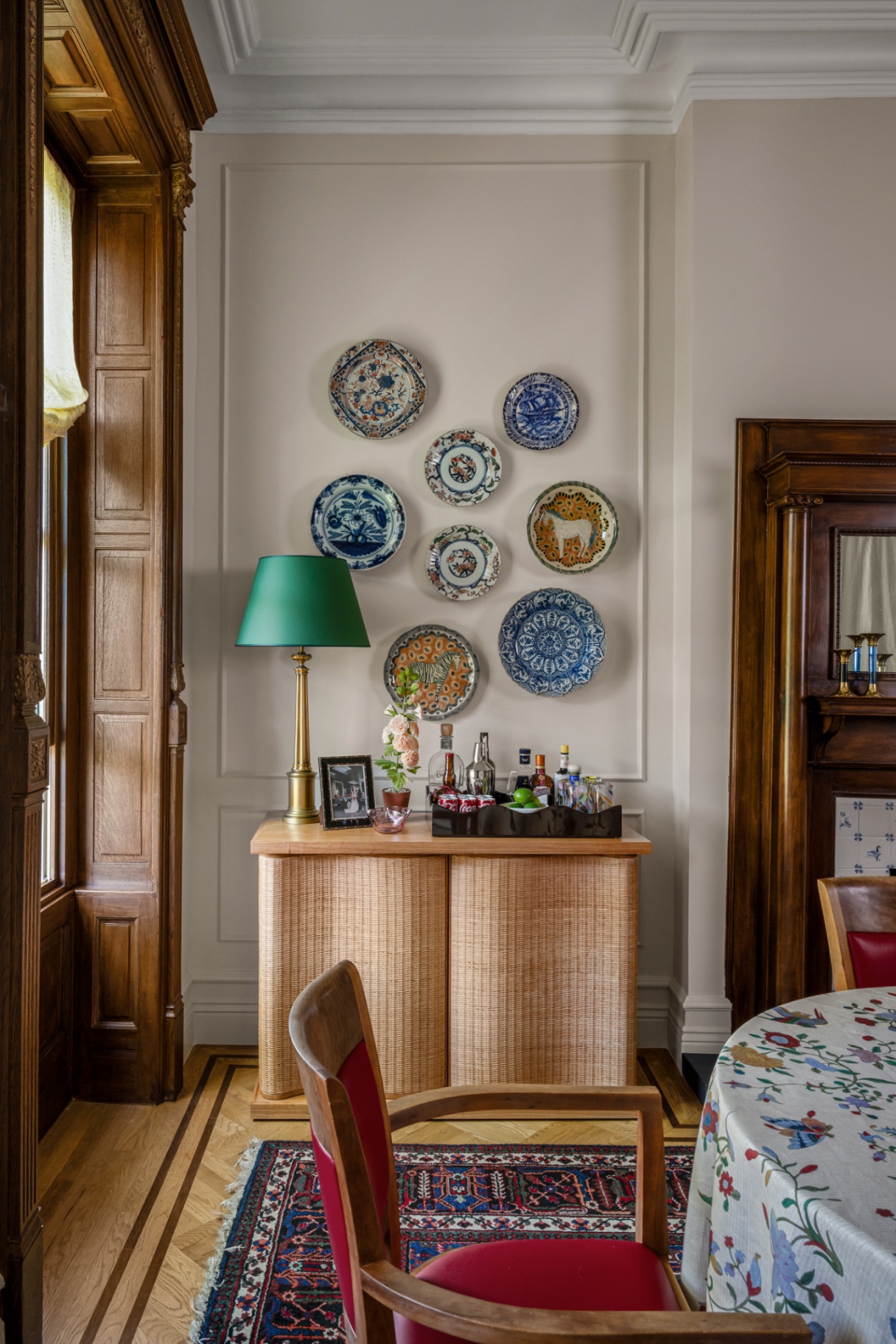 A close-up shot in a dining room with the end of the table and an upholstered wooden chair in the foreground. In the background is a wavy rattan console topped with a table lamp and barware, with plates hanging on the wall above