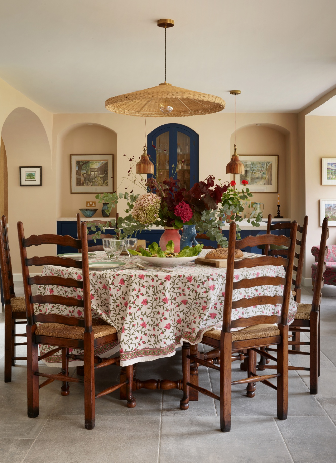 Open-plan dining area with a round wooden table, floral tablecloth, a wide woven central shade, and two hanging accent lights.