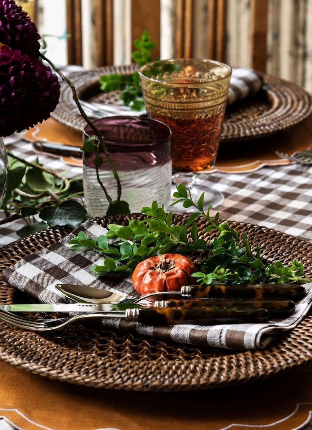 close up of an autumn table with a rattan charger plate with a white and brown linen napkin, a set of tortoiseshell cutlery and a mini pumpkin on top and two tumblers behind it