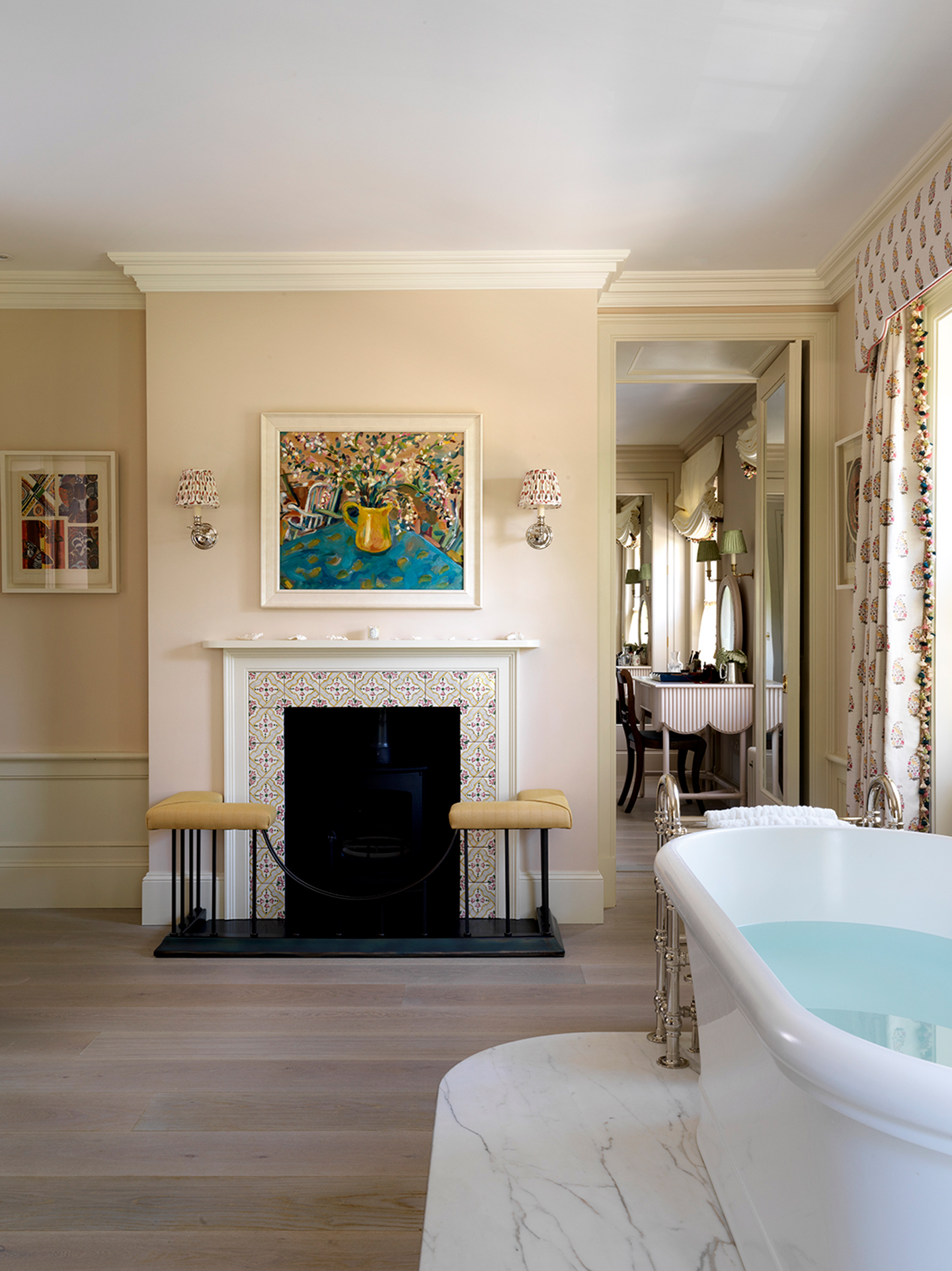A large bathroom with a freestanding bath on a marble platform in the foreground and a fireplace surrounded by tiles and a fender with a large floral artwork and two wall lights hanging above. A doorway to the right leads through to a dressing room