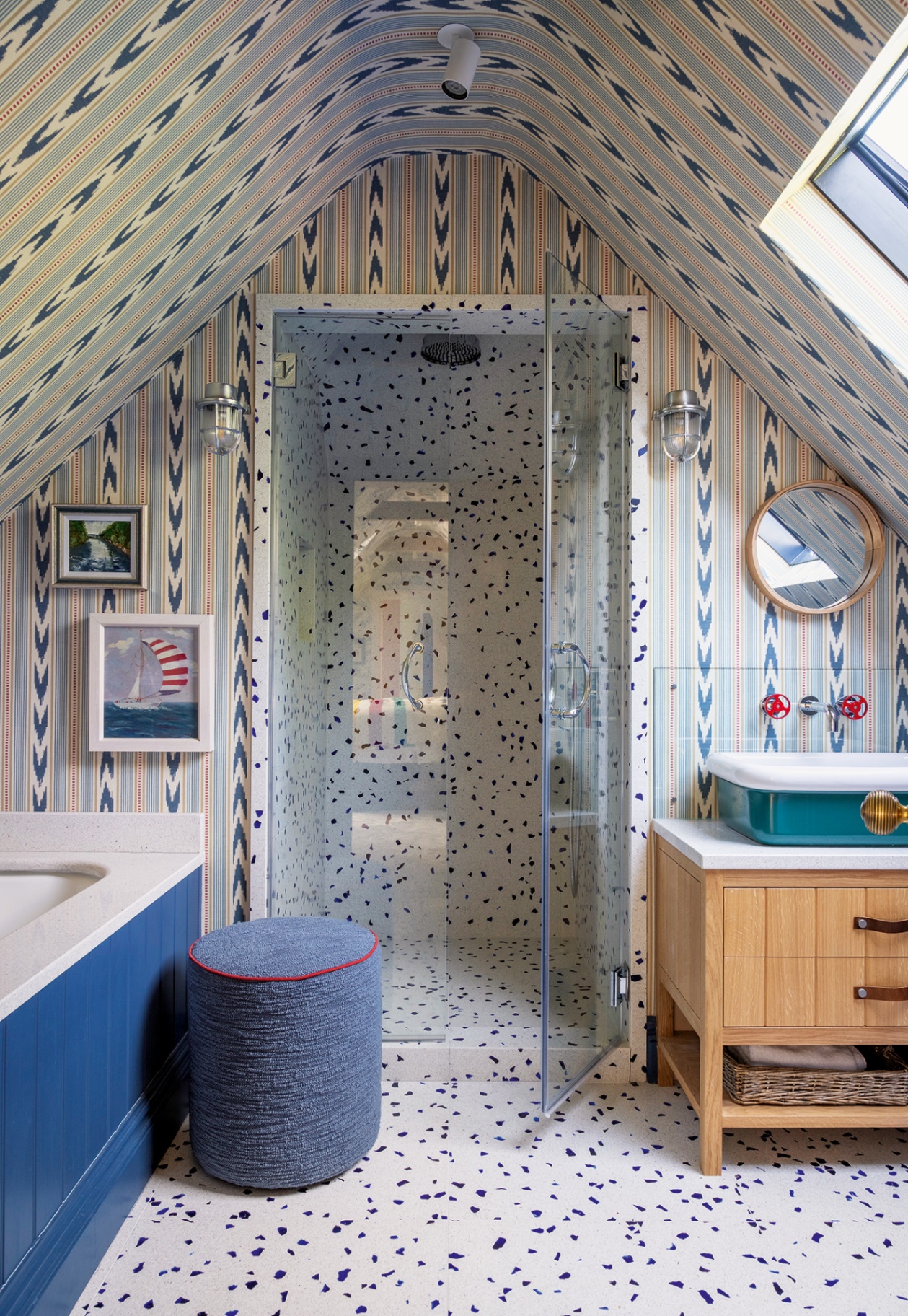 A bathroom with a sloped curved ceiling. The floor is made of blue and white terrazzo tiles and the walls are covered in an ikat stripe paper. A shower is built into the back wall, with a bath to the left and a vanity unit to the right