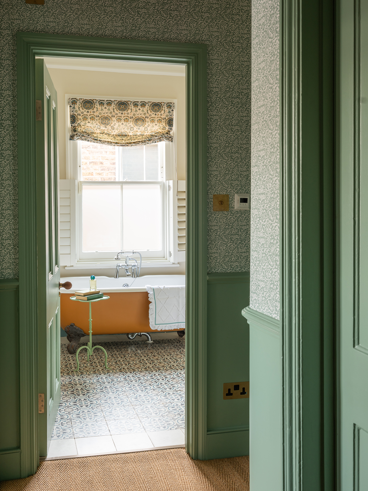 A hallway with the lower half of walls painted green and the upper half in green wallpaper. Through an open door a bathroom can be seen with a yellow freestanding bath and patterned blinds
