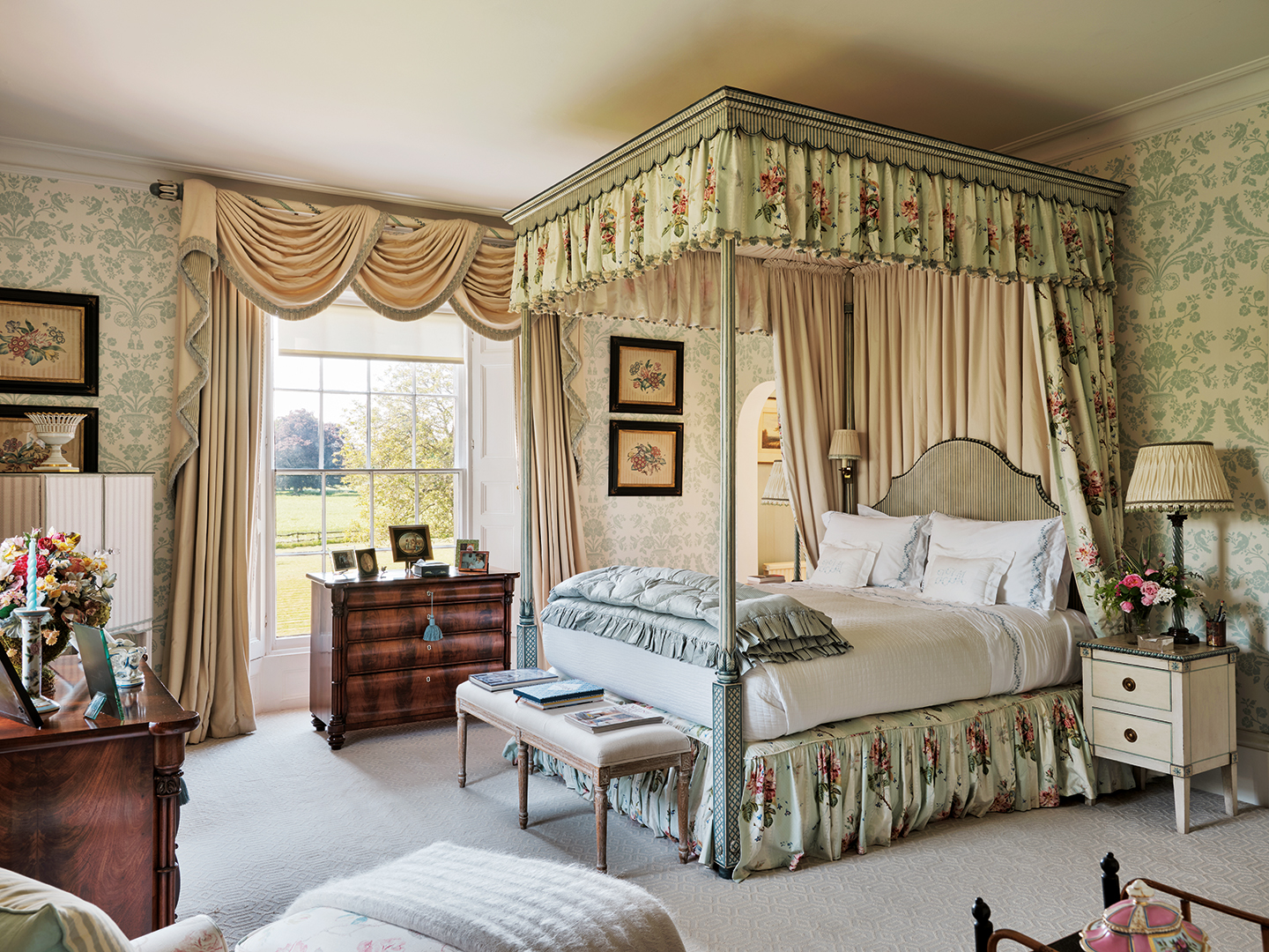 a grand bedroom with a double four poster bed with floral curtains and valance, dressed in white bed linen and a folded blue eiderdown, a bench sits at the end of the bed and a chest of drawers in front of the sash window dressed with ornate curtains