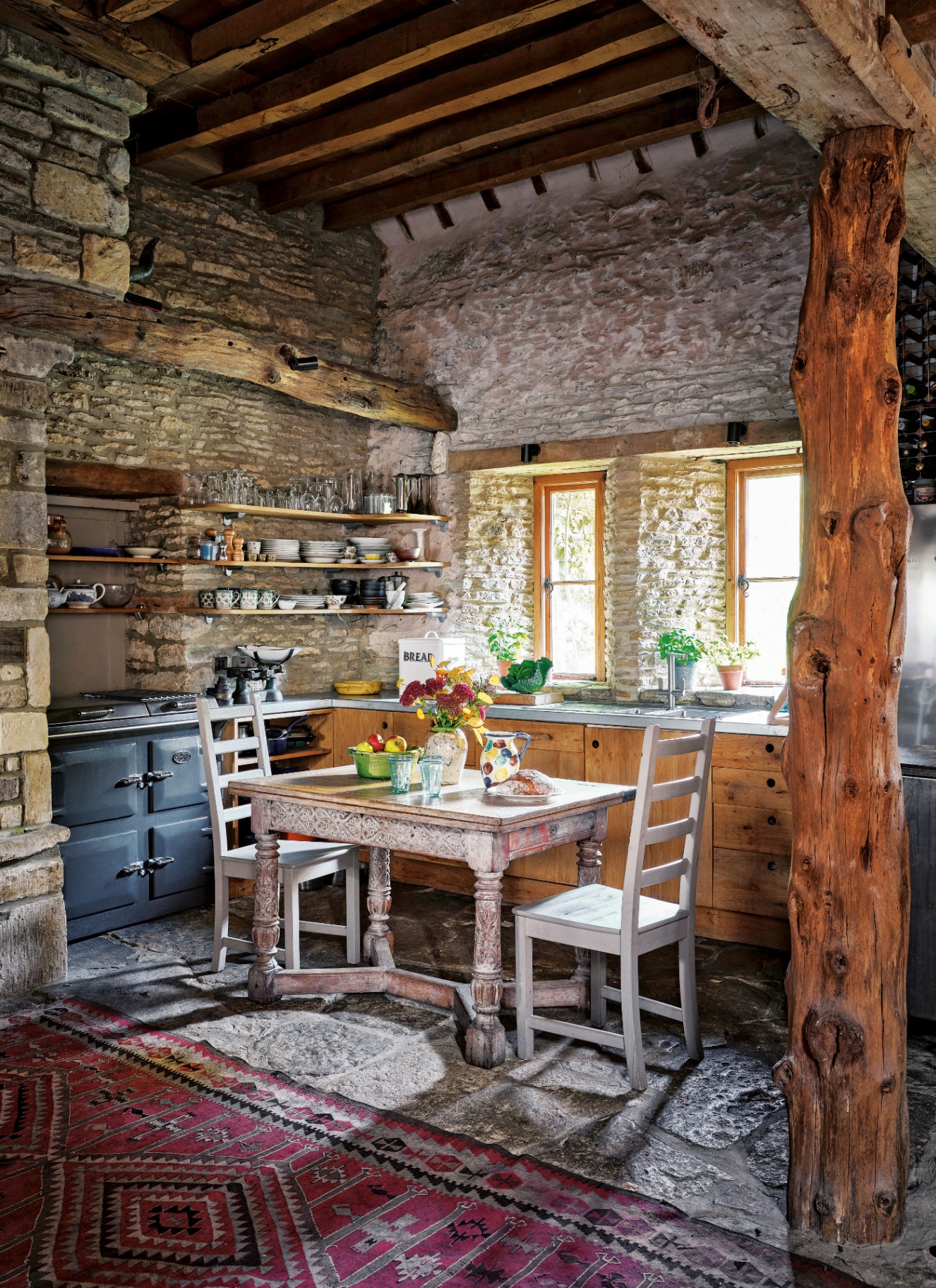 a kitchen with exposed brick walls, wooden beams, a small wooden table and chairs, a grey aga and open shelves filled with plates and glasses