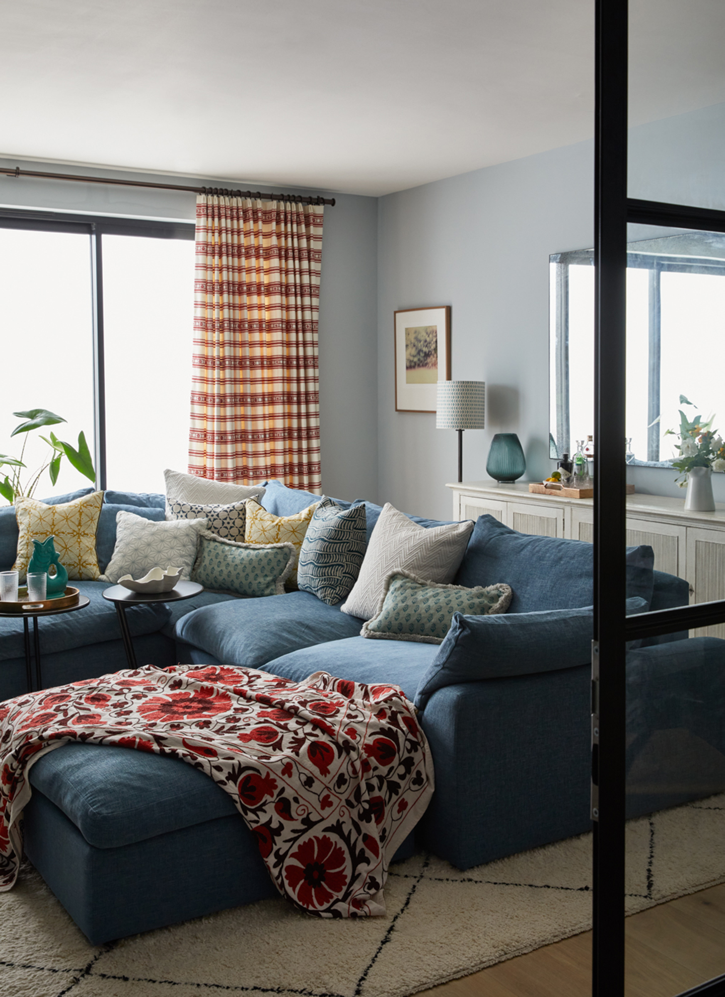 A modern living room with pale blue walls, a deep blue corner sofa dressed with patterned cushions and a red floral throw, patterned curtains, and glass patio doors