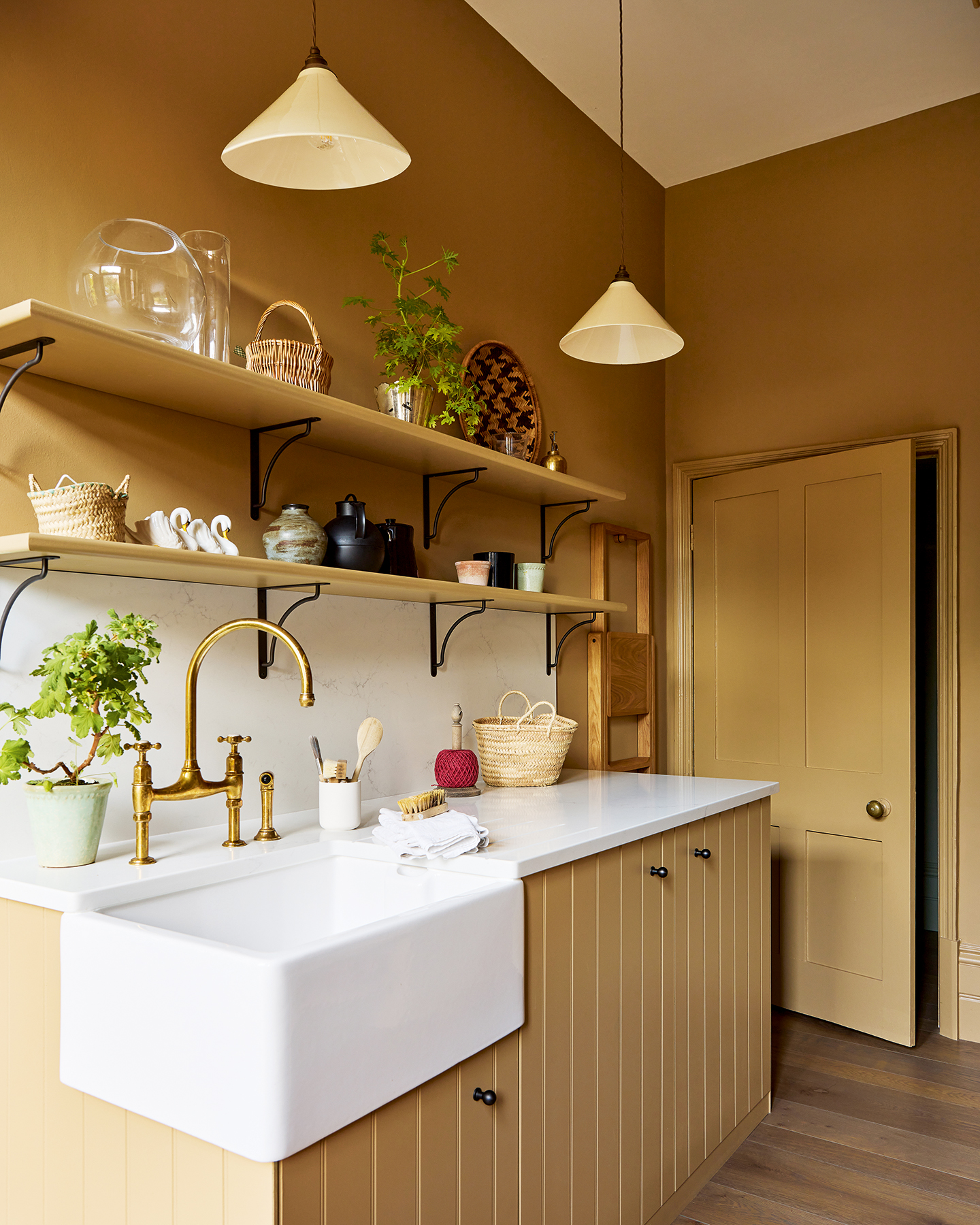A utility room with ochre walls, woodwork and joinery. Tongue and groove cabinetry features a built-in belfast sink and white marble worktops and backsplash, above are two open shelves holding baskets, vases and plants, and two ceramic lights hang above.