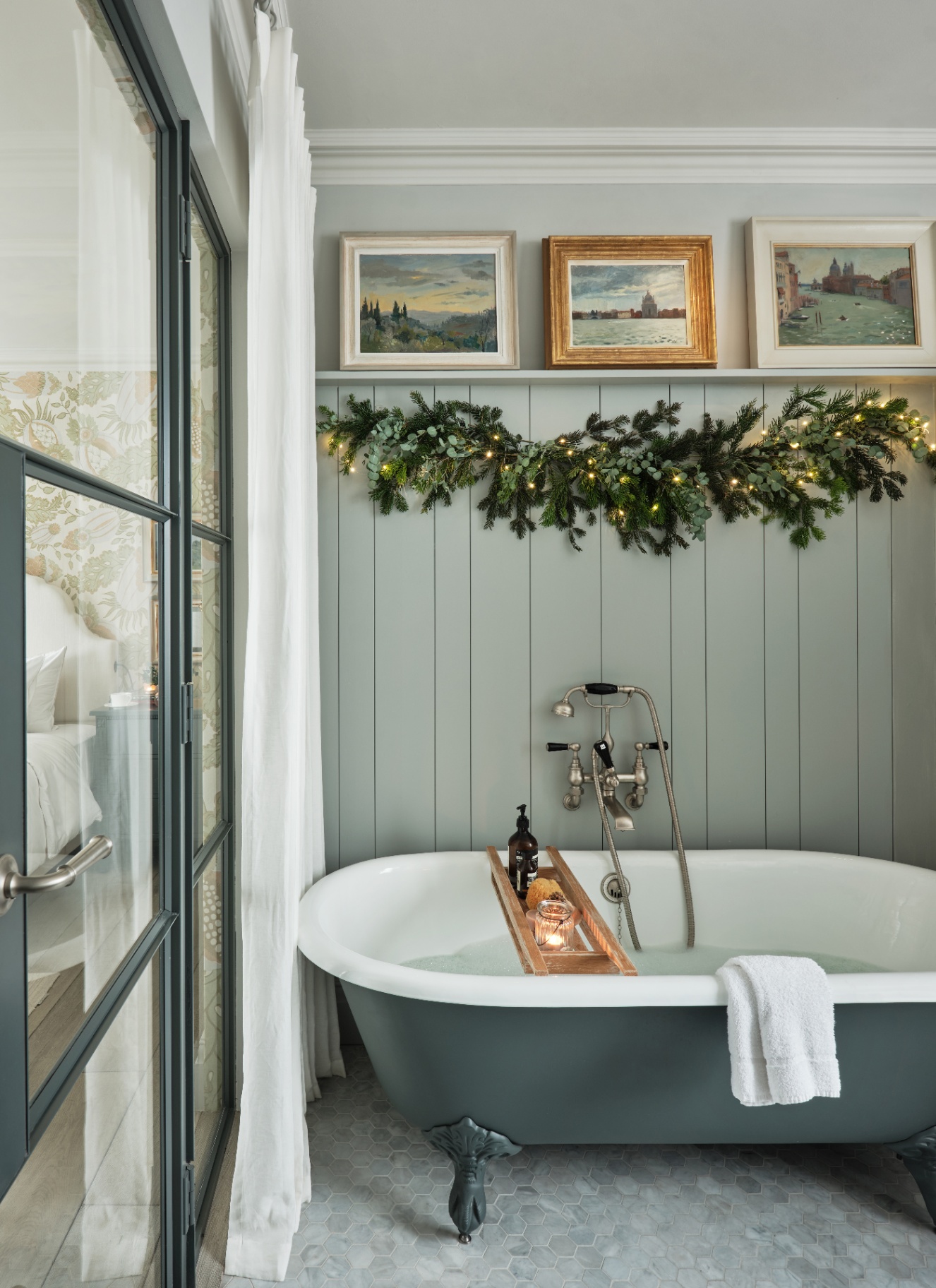 Luxury bathroom with a clawfoot bath, wooden tub caddy, and a festive evergreen garland with string lights mounted on the wall.