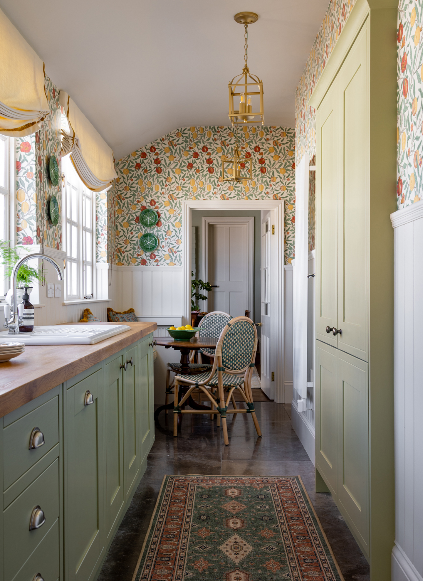 a narrow kitchen with sage green cabinetry, a patterned runner, a small round dining table and chairs and red, white, green and yellow patterned wallpaper