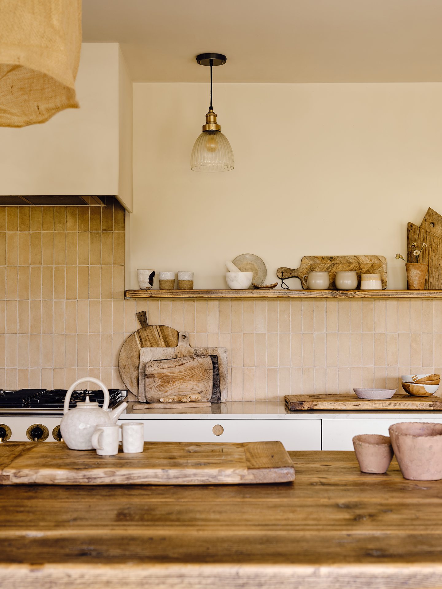 A close up shot of a kitchen with the wooden top of an island in the foreground, topped with a large chopping board and a kettle and cups, while a tiled backsplash lines the back wall along with a freestanding shelf holding kitchenware