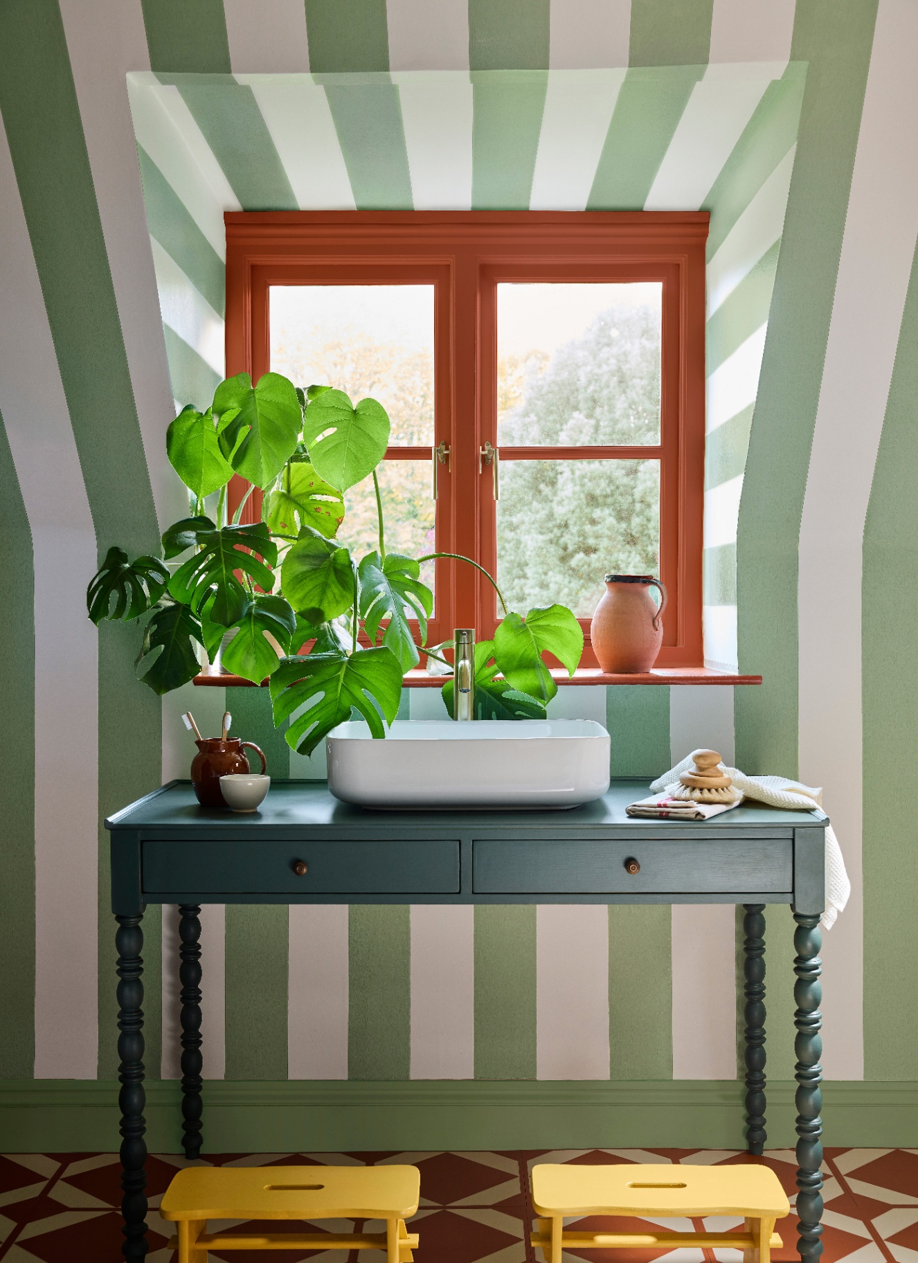 a room with sloped ceiling painted in green and white stripes and a dark green bobbin table in front of a window with a plant on top