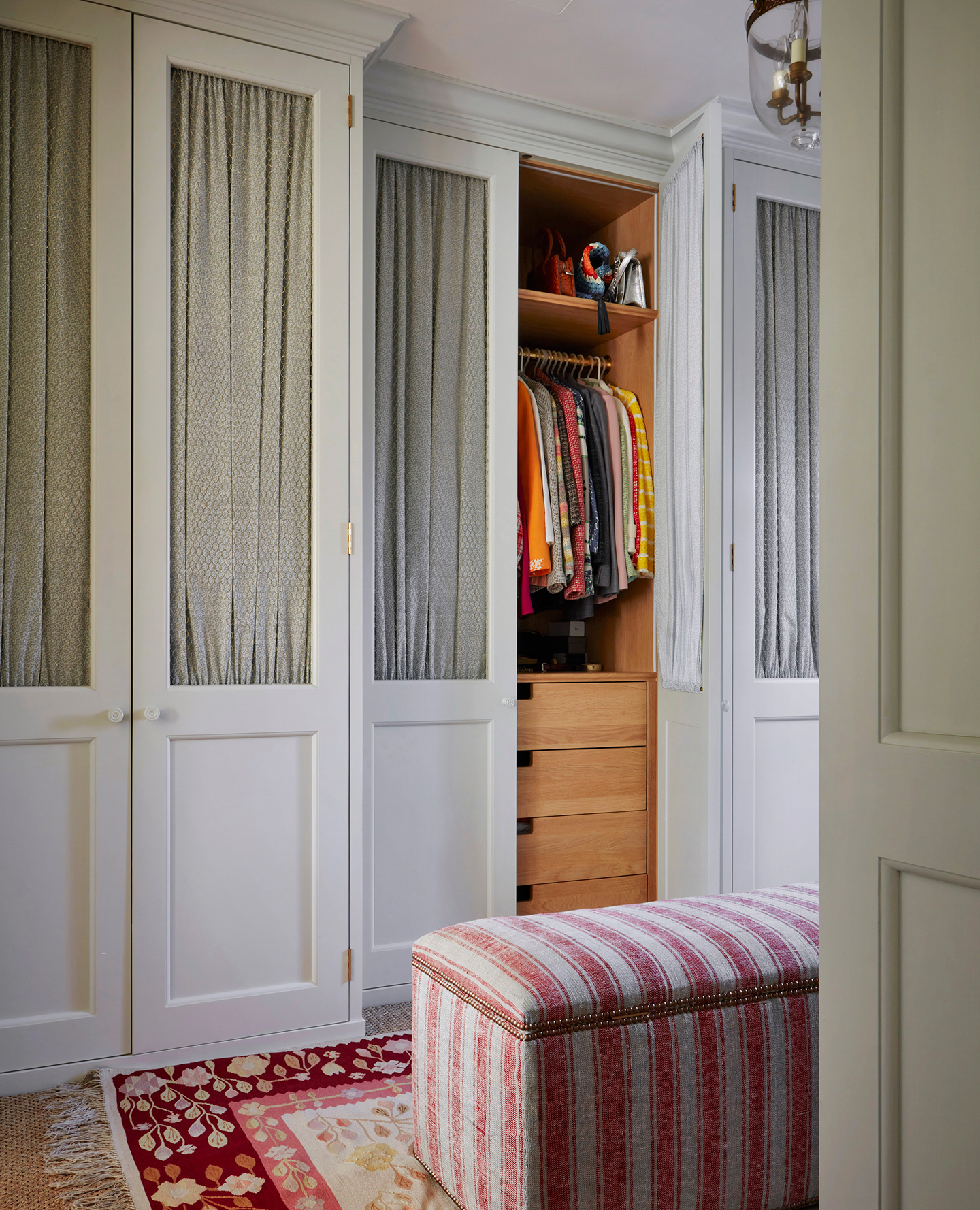 A small dressing room with a wall of built-in wardrobes with fabric panel fronts and in the foreground is a striped storage bench in a red and white fabric placed on top of a red, pink and white rug