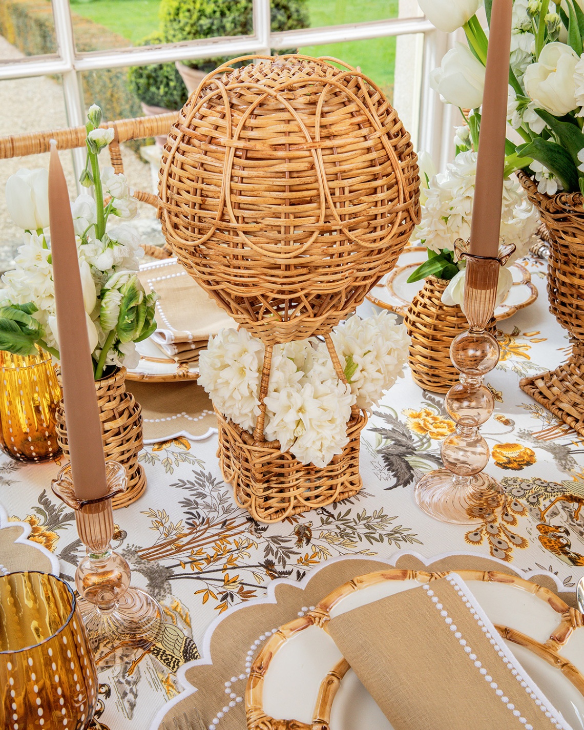 a spring tablescape with white and beige floral tablecloth, plates and candle sticks and a large rattan hot air balloon centrepiece