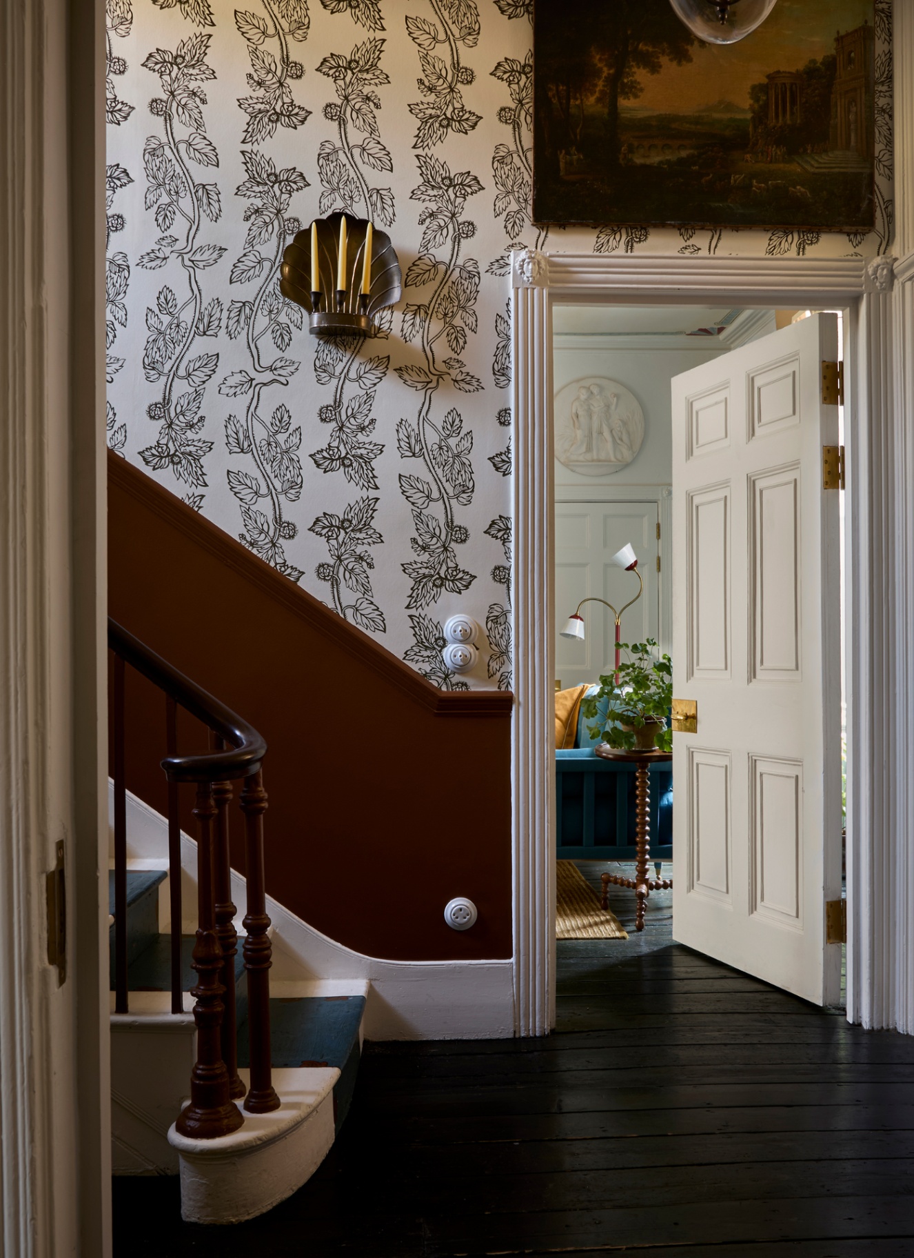 a hallway with white wallpaper with black leaf illustrations, a dado rail panelling painted in dark orange, a shell shaped candle sconce with 3 tapered candles, a white open door and dark wooden flooring