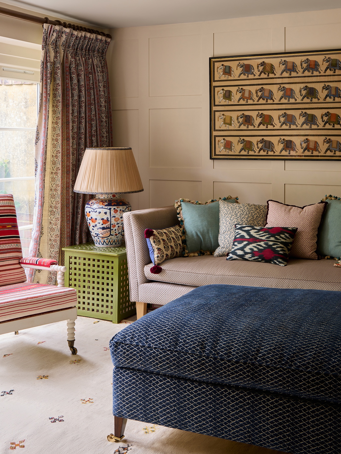 A light-filled living room with neutral, panelled walls. A sofa topped with colourful cushions and a striped armchair are arranged around an ottoman. A side table holds a large lamp. There is an elephant artwork on the back wall. 