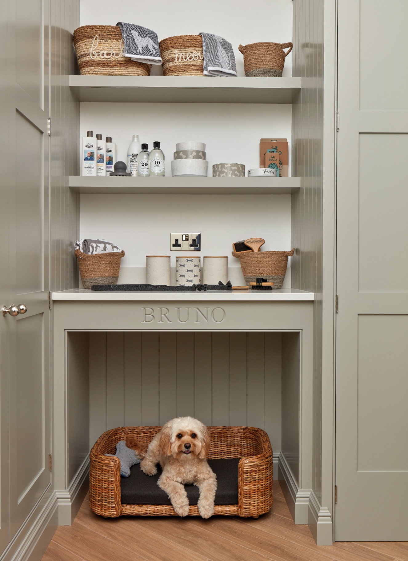 a dedicated dog corner in a utility room with grey cabinetry, a dog bed alcove with a wicker bed and a dog inside with shelves above it filled with dog accessories