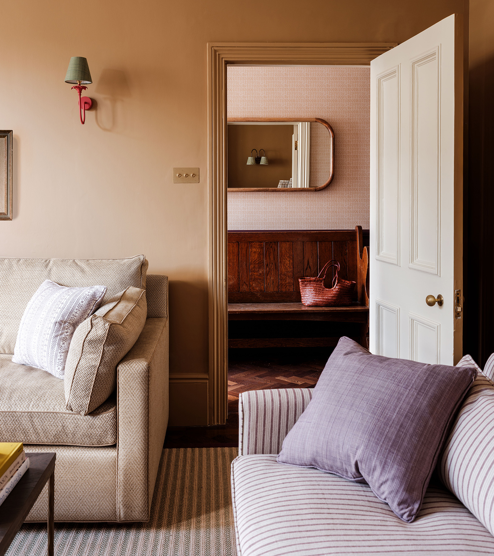 A living room corner with beige walls and skirting board, with two corners of beige sofas, a red sconce on the wall with the door open looking through to a dark bench in the hallway