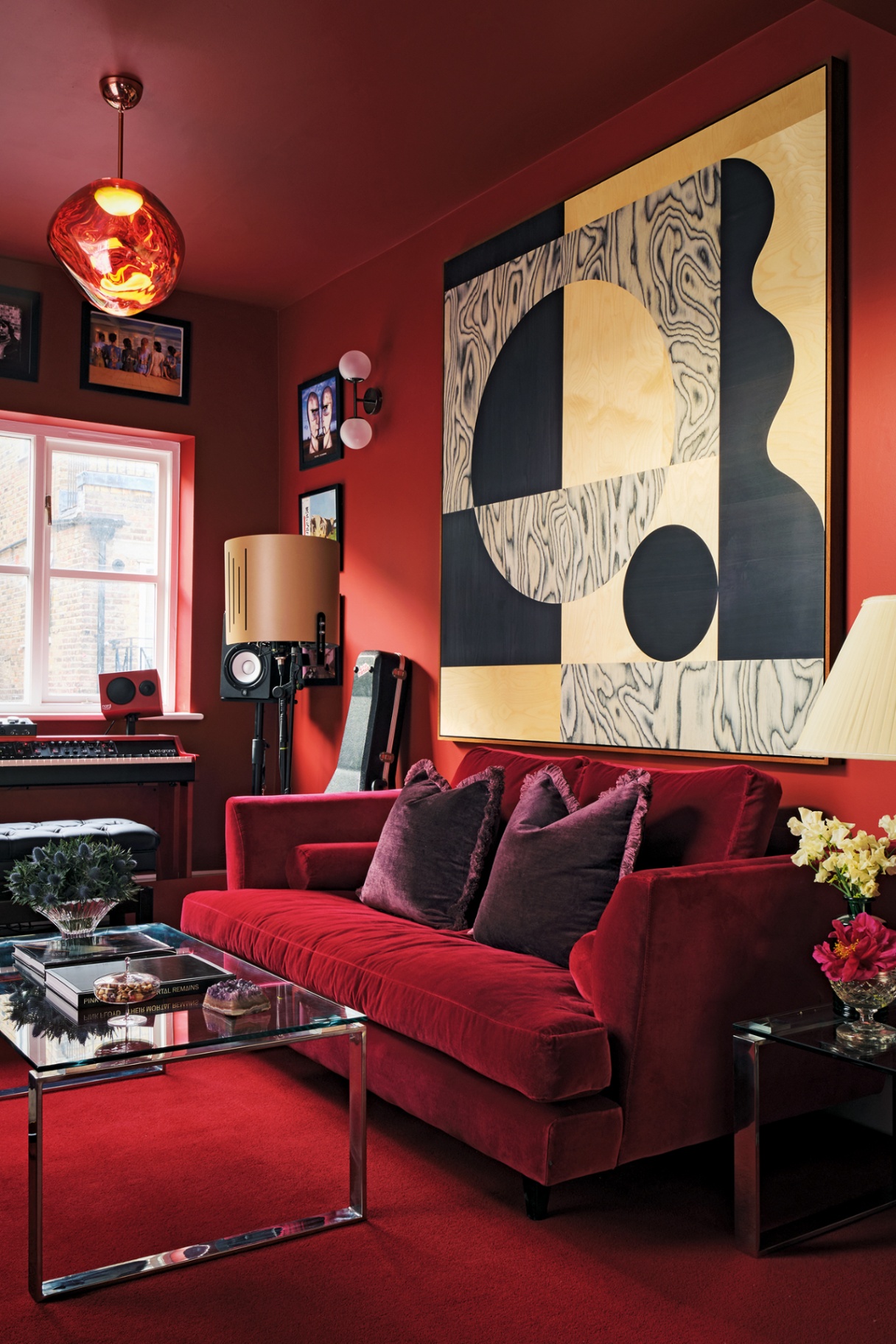 a music room with red walls and carpet, and a red sofa dressed with purple velvet cushions. A keyboard, guitar and speaker are arranged around the room. A glass and chrome coffee table sits to the left. On the wall a large abstract artwork is displayed