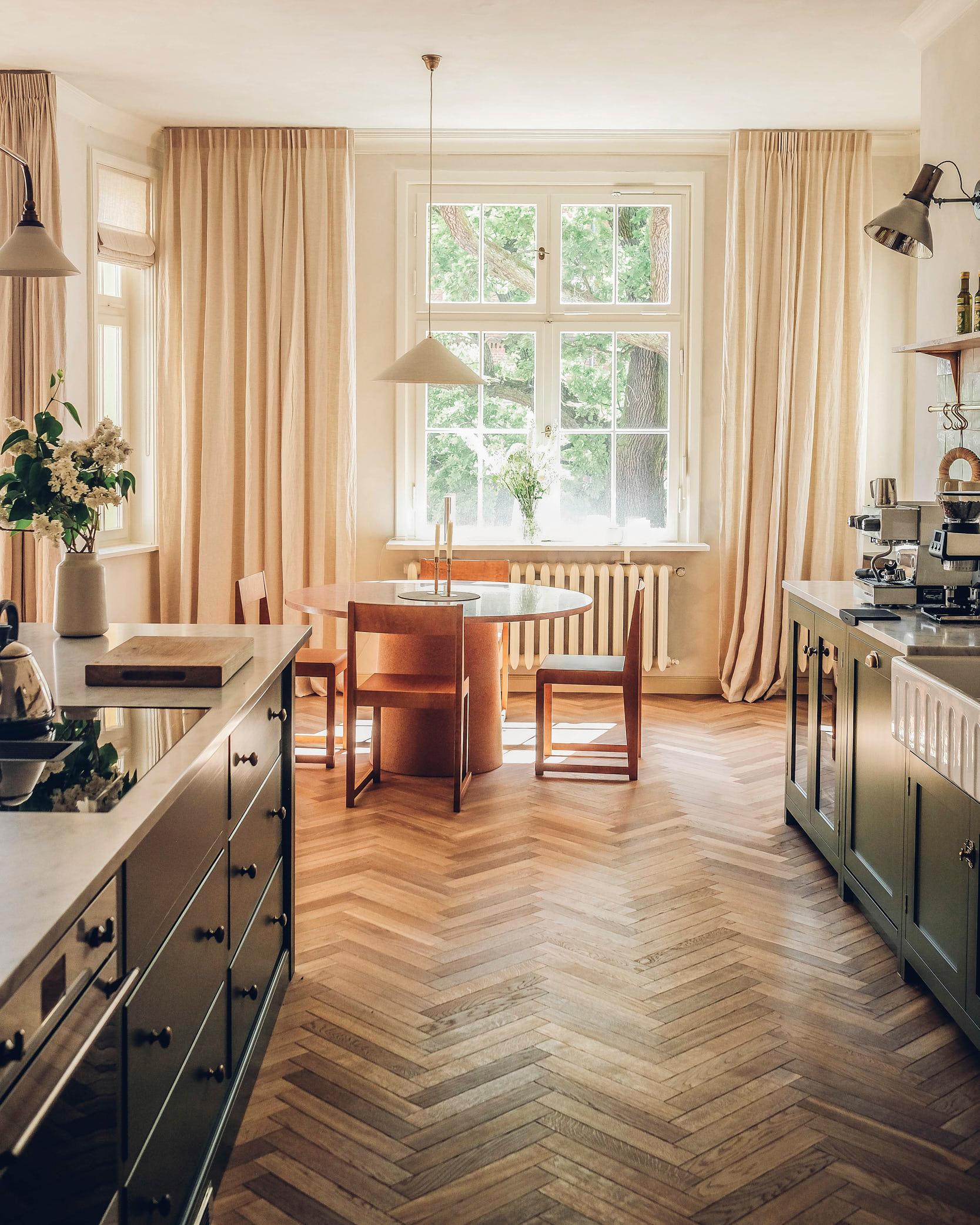 a kitchen with wooden parquet flooring, an round orange dining table with 4 matching seats and a white pendant light hanging above it and dark green kitchen cabinetry