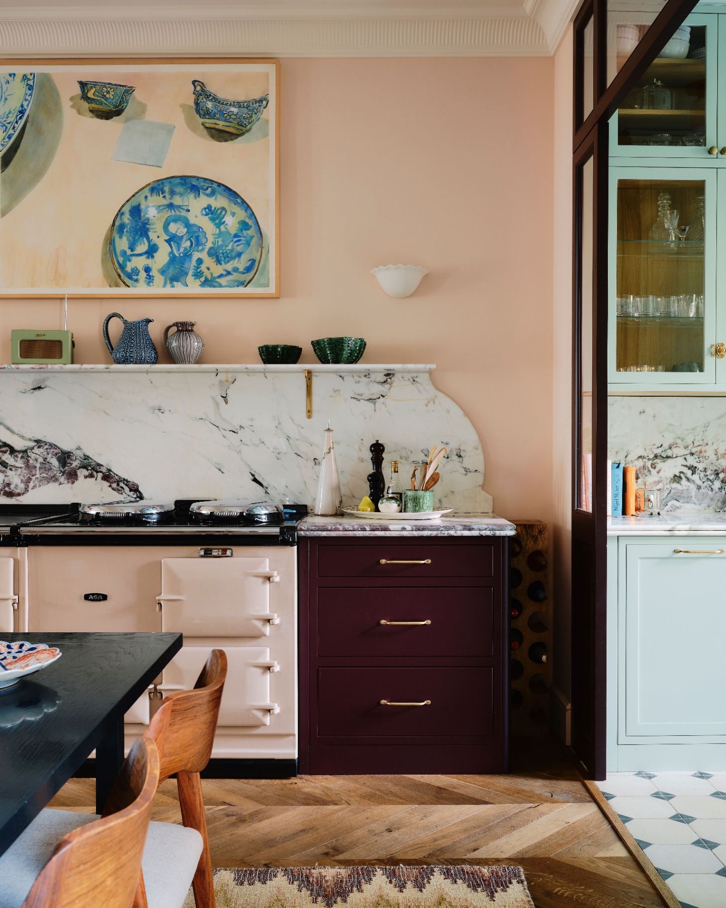 a kitchen with plaster pink walls, a white marble splashback, a pale pink range cooker, a cabinet painted in burgundy and a large painting of blue plates