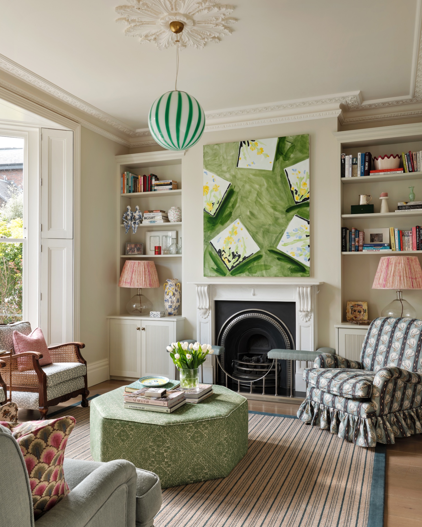 a light and bright living room with a statement white and green stripe pendant, alcove shelving units either side of a fireplace with a large green print above it, an upholstered green ottoman and a blue stripe skirted armchair